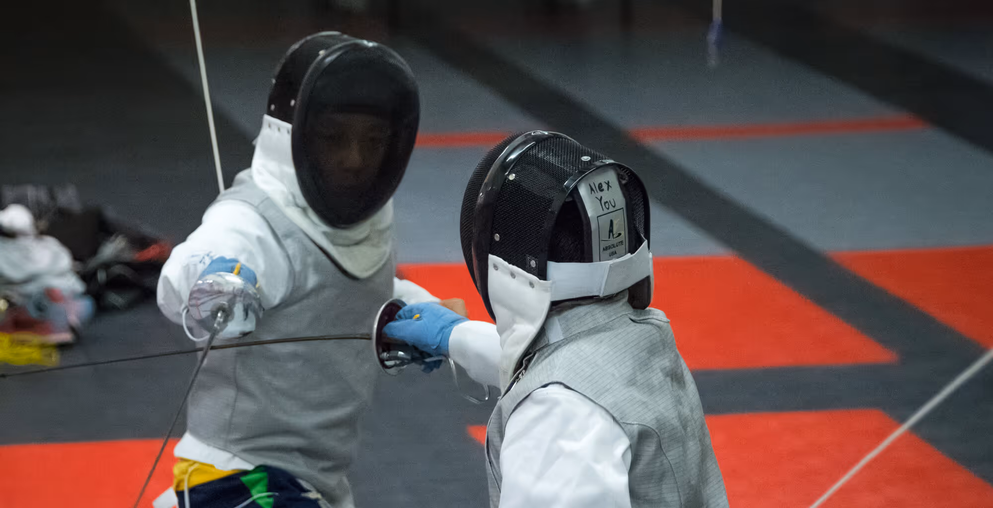 Two fencers in protective gear engaged in a match on a red and gray floor.