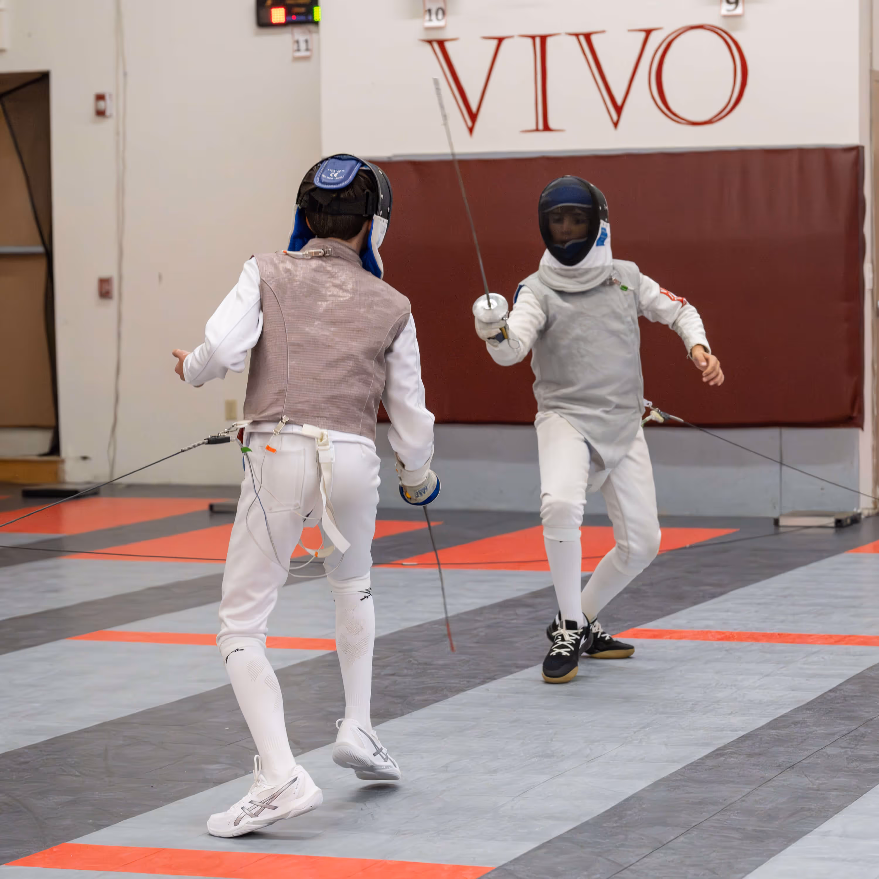 Two young fencers in protective gear engaged in a match on a marked floor.
