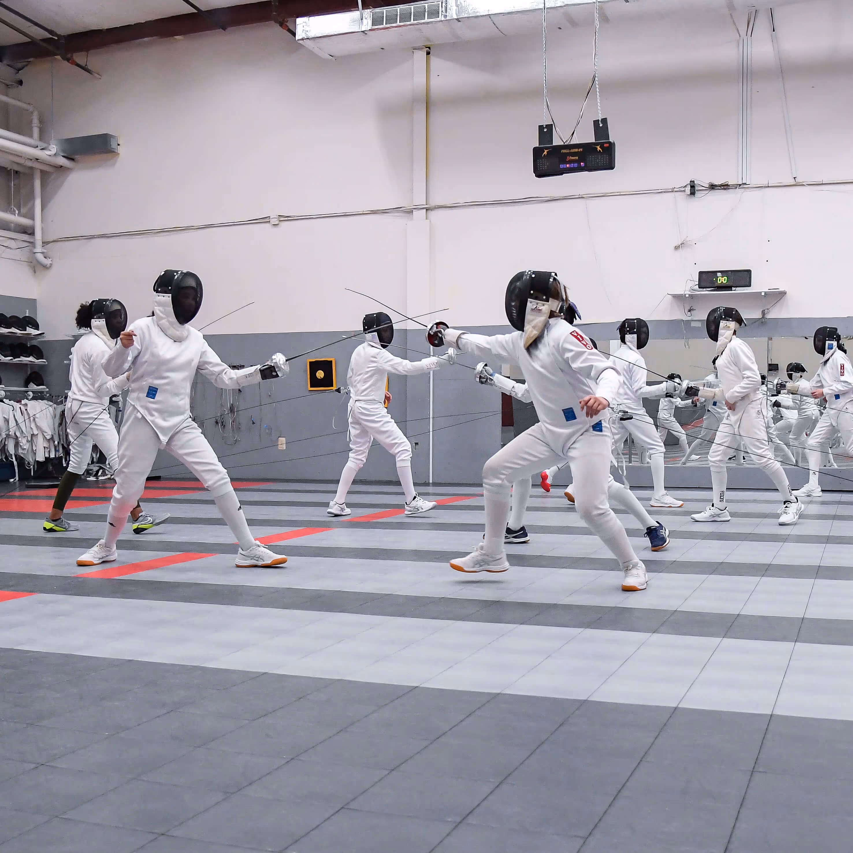Group of fencers in white uniforms and helmets practicing lunges and sword strikes in a spacious indoor fencing gym.