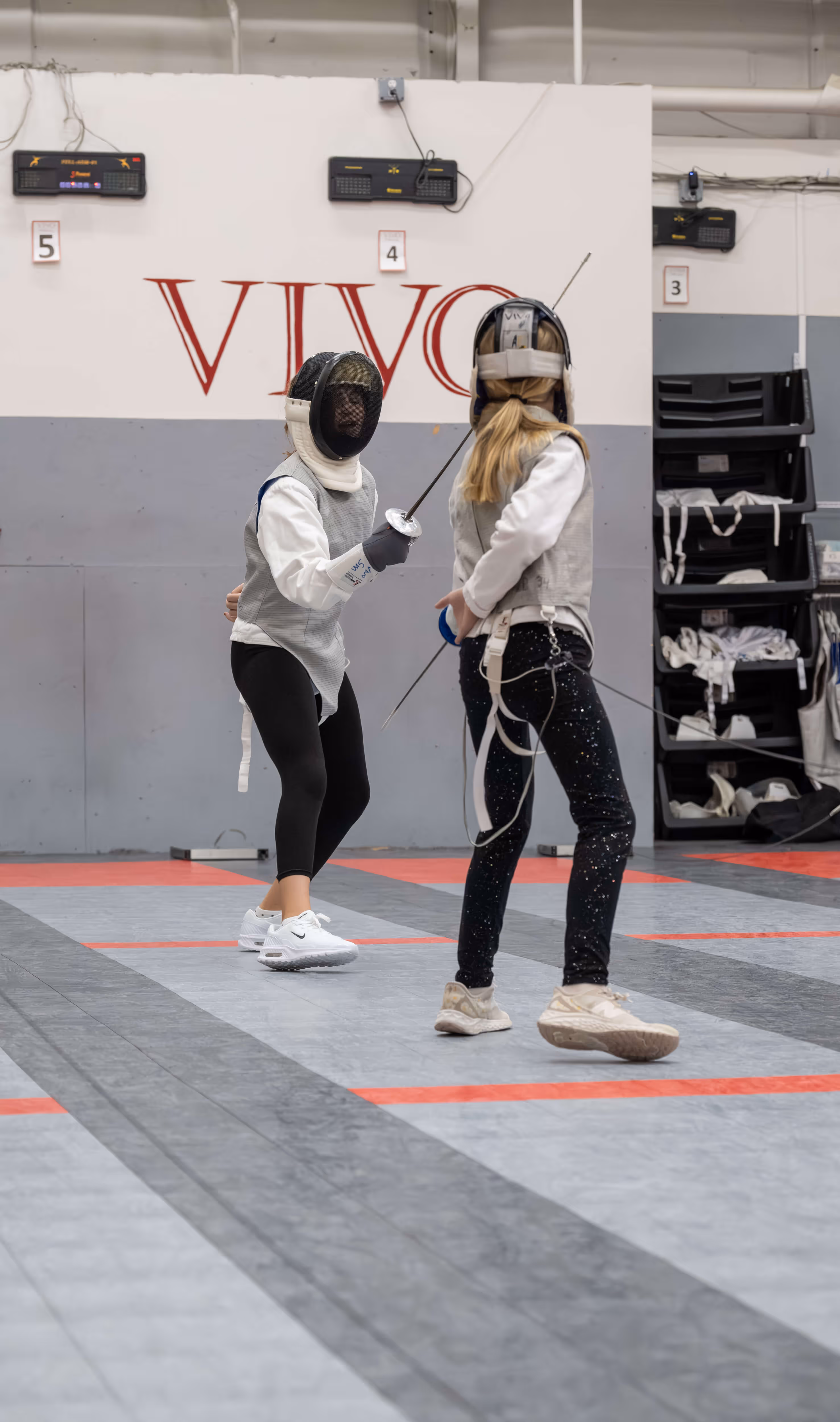 Two young women fencing indoors, wearing protective gear and holding foils on a marked floor.