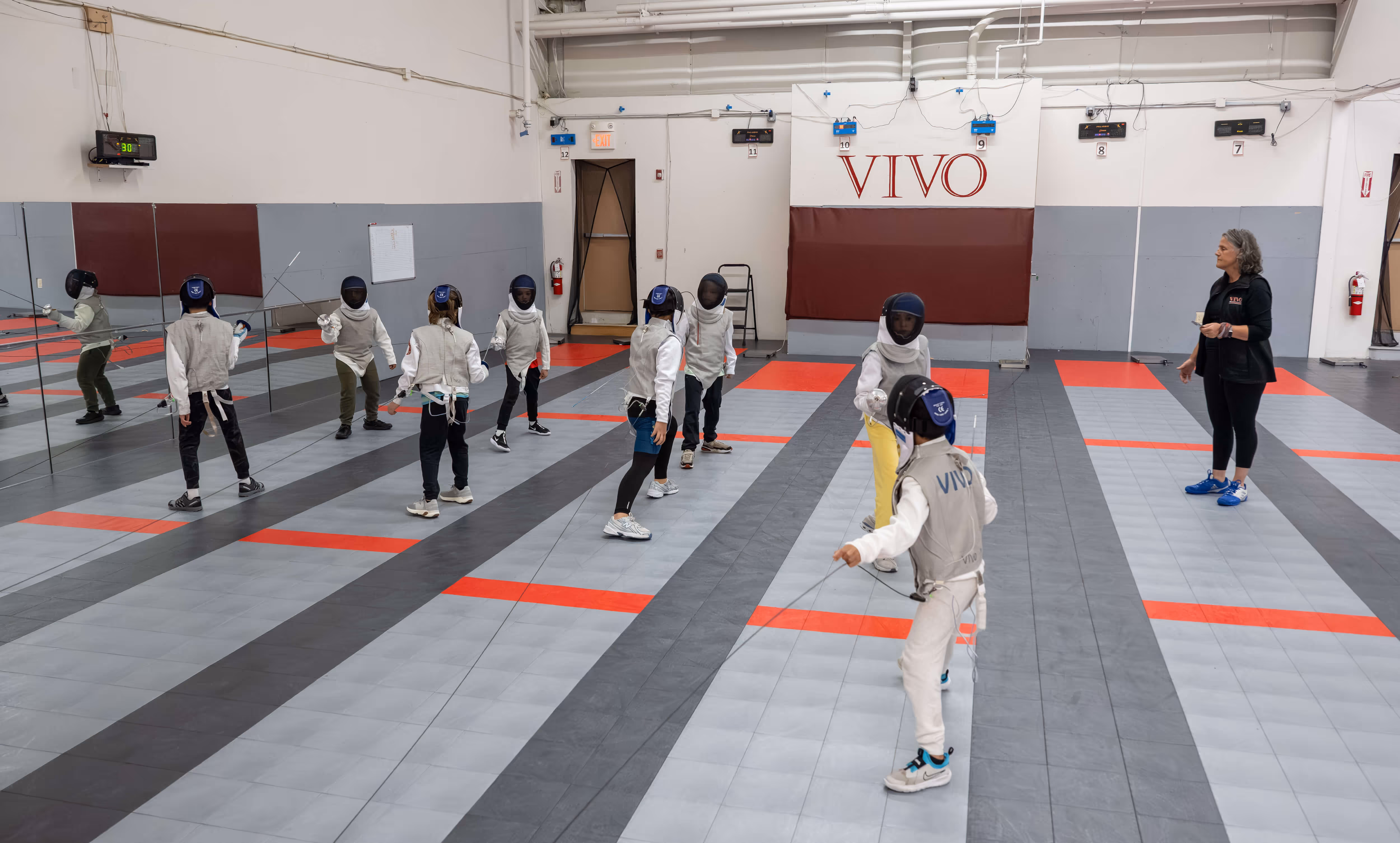 Children in protective fencing gear practicing with foils in a gym, supervised by a coach.