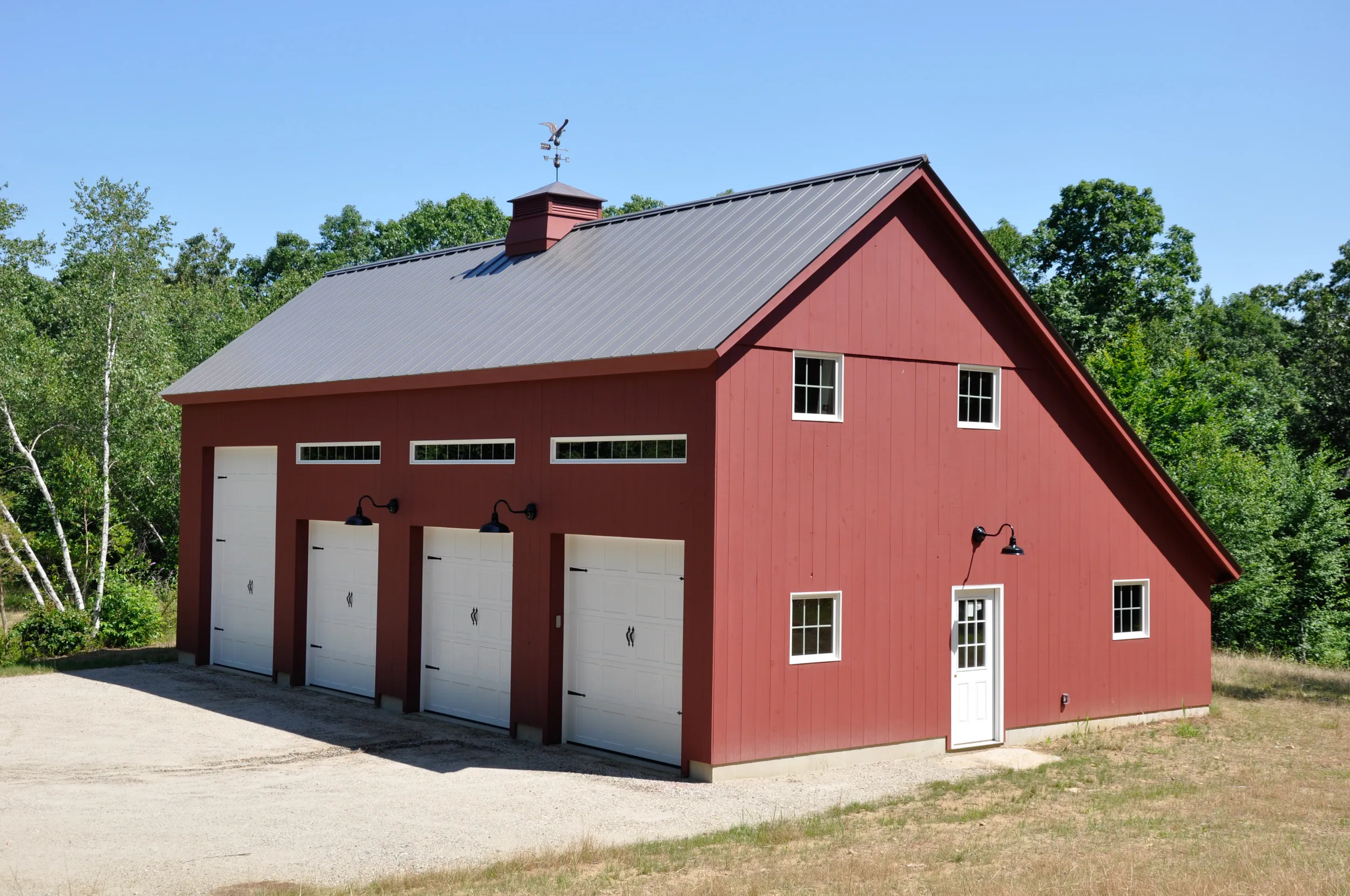 Red barn with four white garage doors, metal roof, and surrounding trees on gravel driveway.