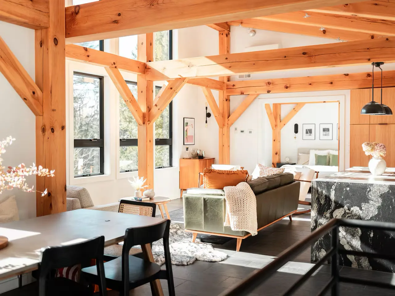 Modern open-plan living space with exposed wooden beams, large windows, contemporary furniture, and pendant lighting.