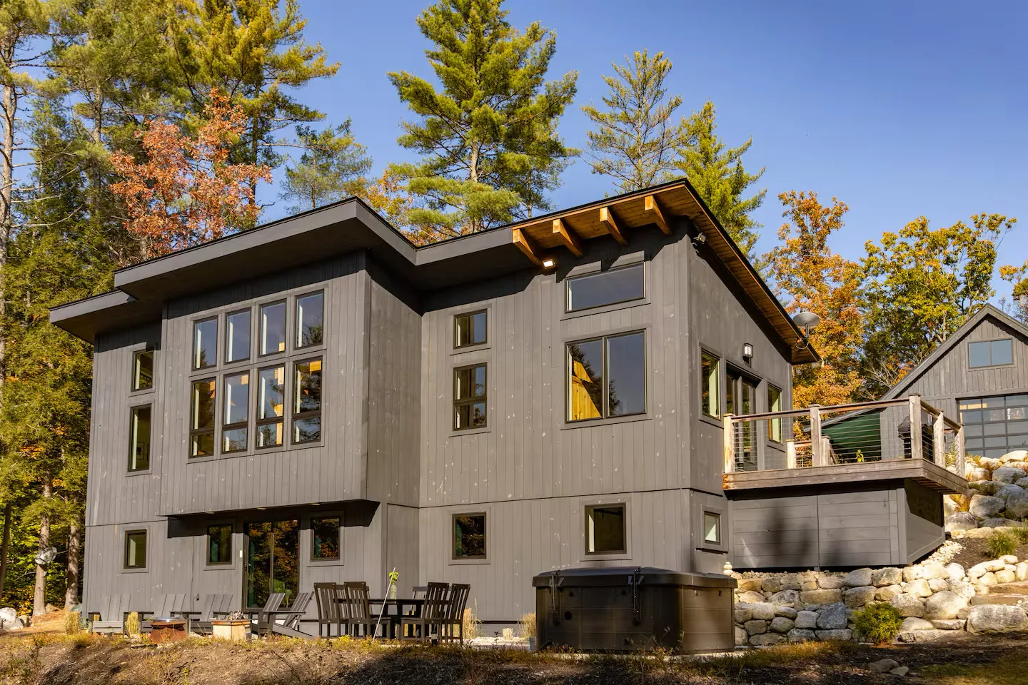 Modern gray house with large windows surrounded by autumn trees and stone foundation.