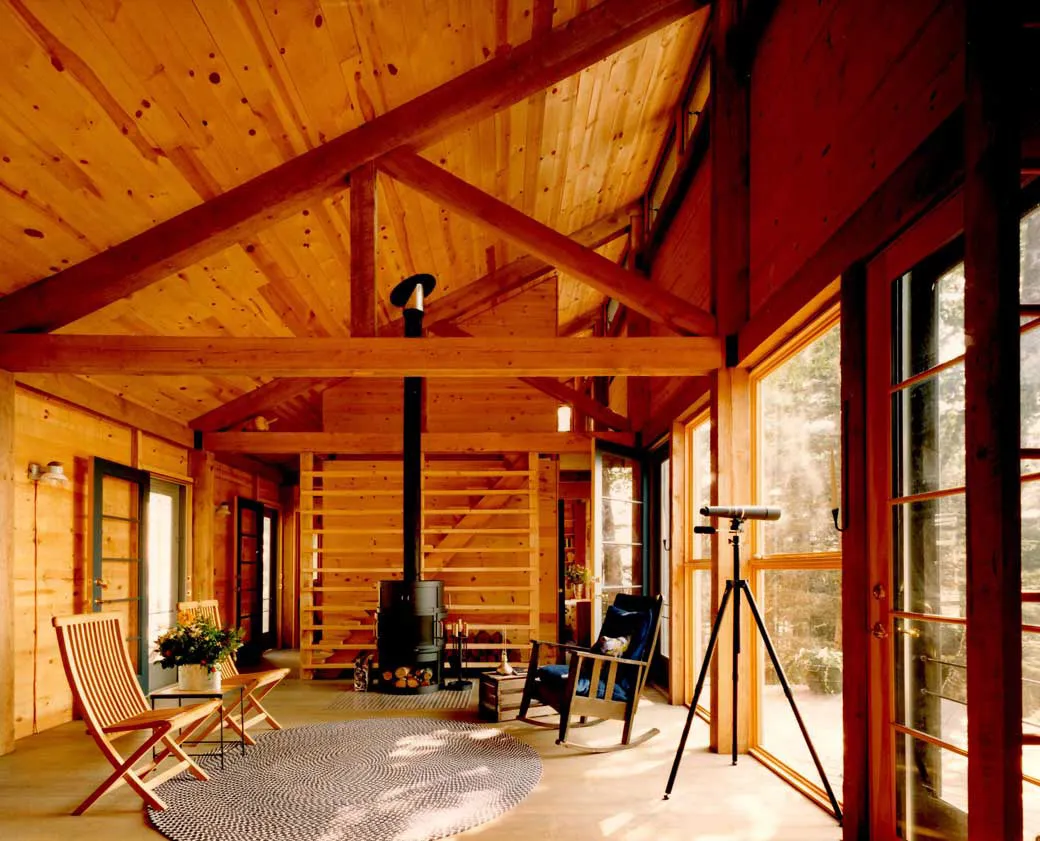 Modern wooden cabin interior with wood stove, large windows, and minimalist furniture.