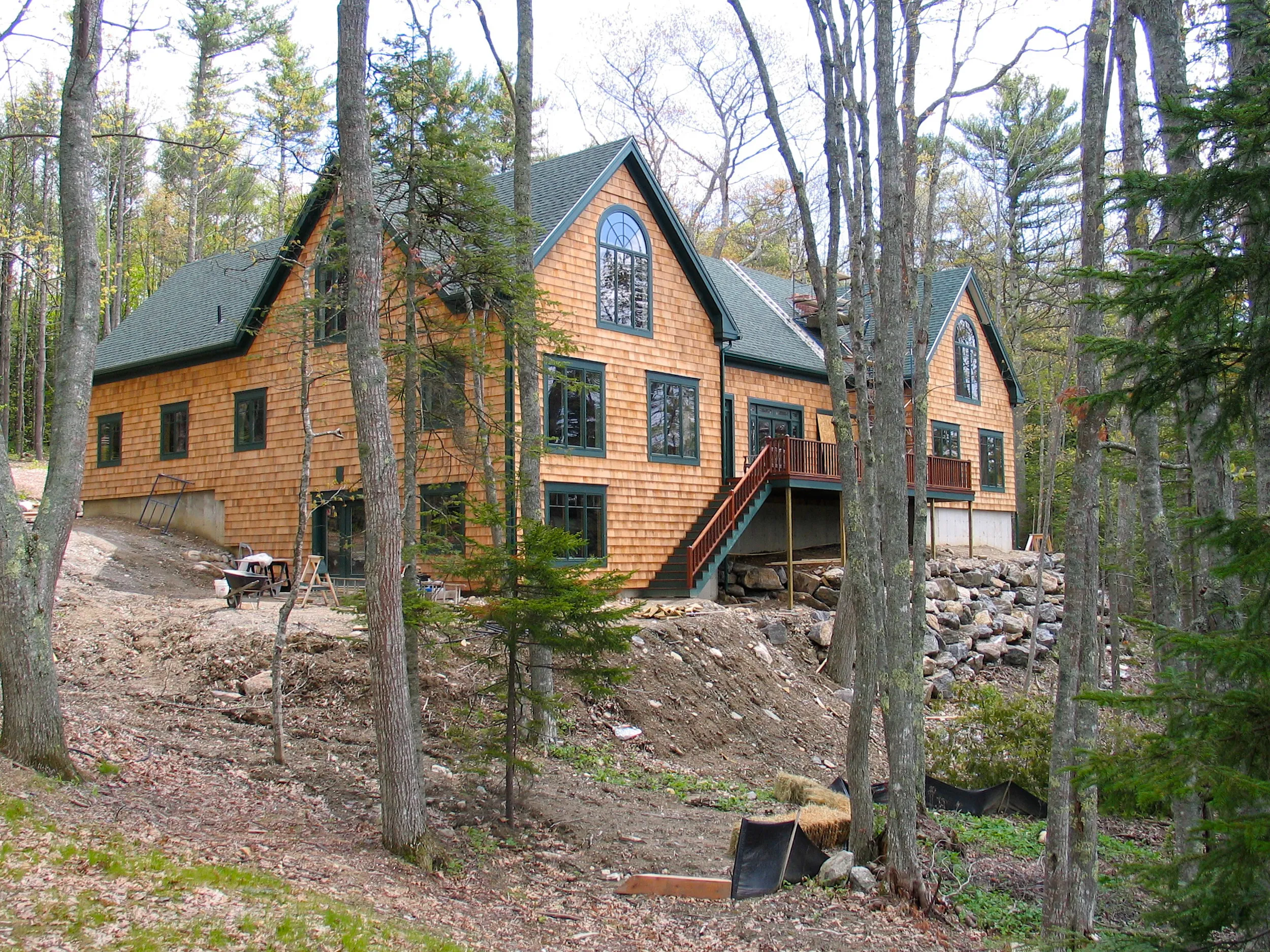 Modern orange wood-sided house with dark roofs nestled among tall forest trees and rocky terrain.