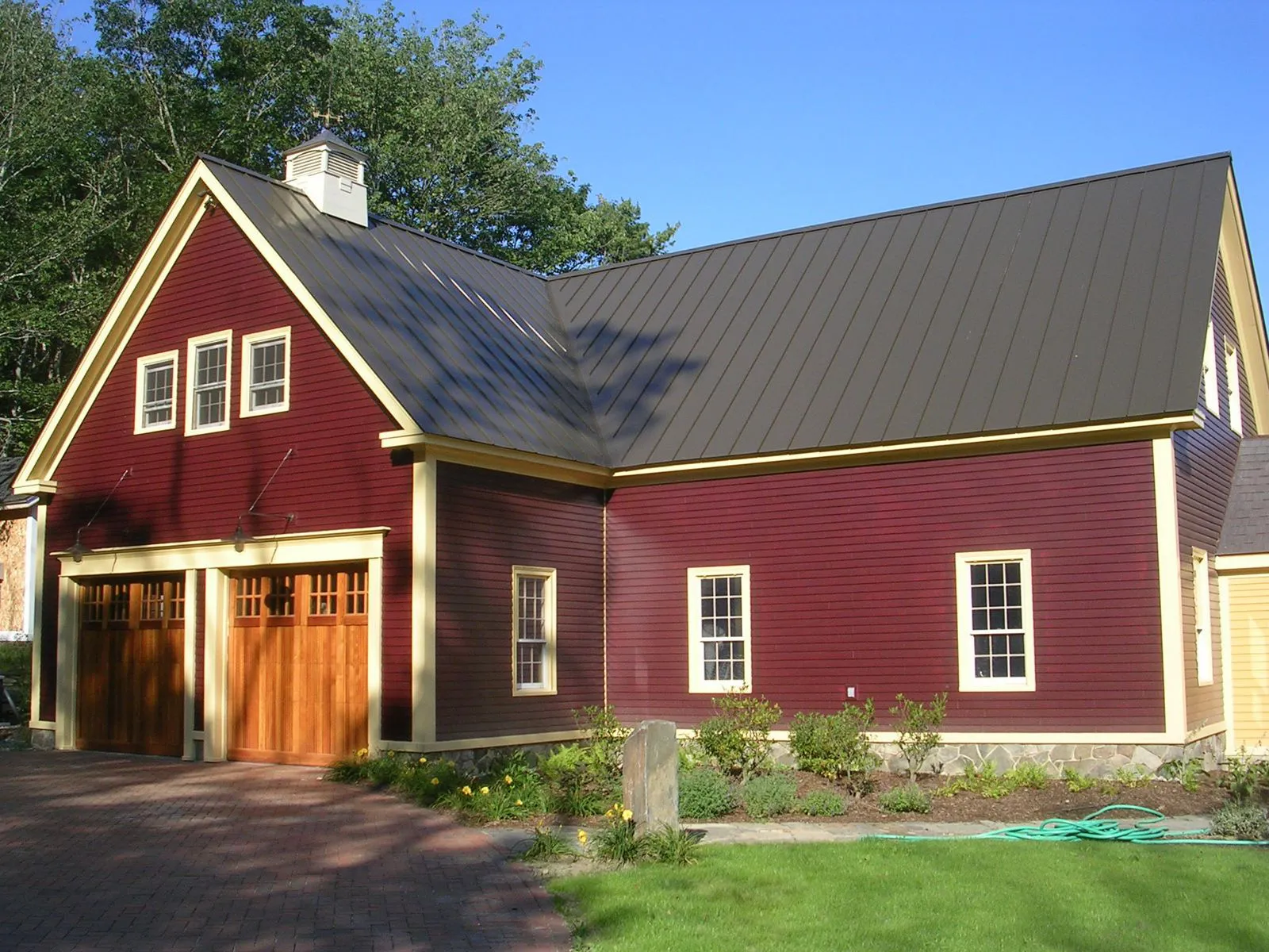 Red farmhouse with metal roof, wooden garage doors, and white trim surrounding green landscaping