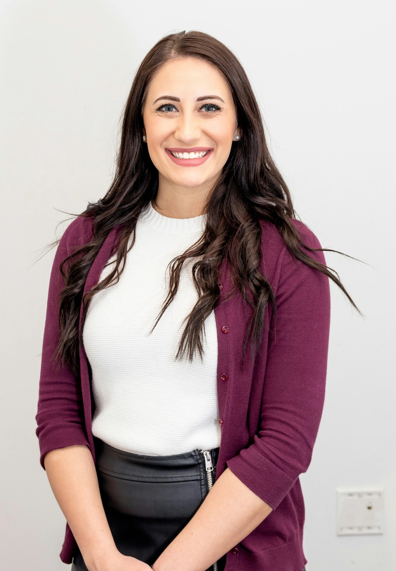 Smiling woman with long dark hair wearing a white sweater, purple cardigan, and black skirt against a plain background.
