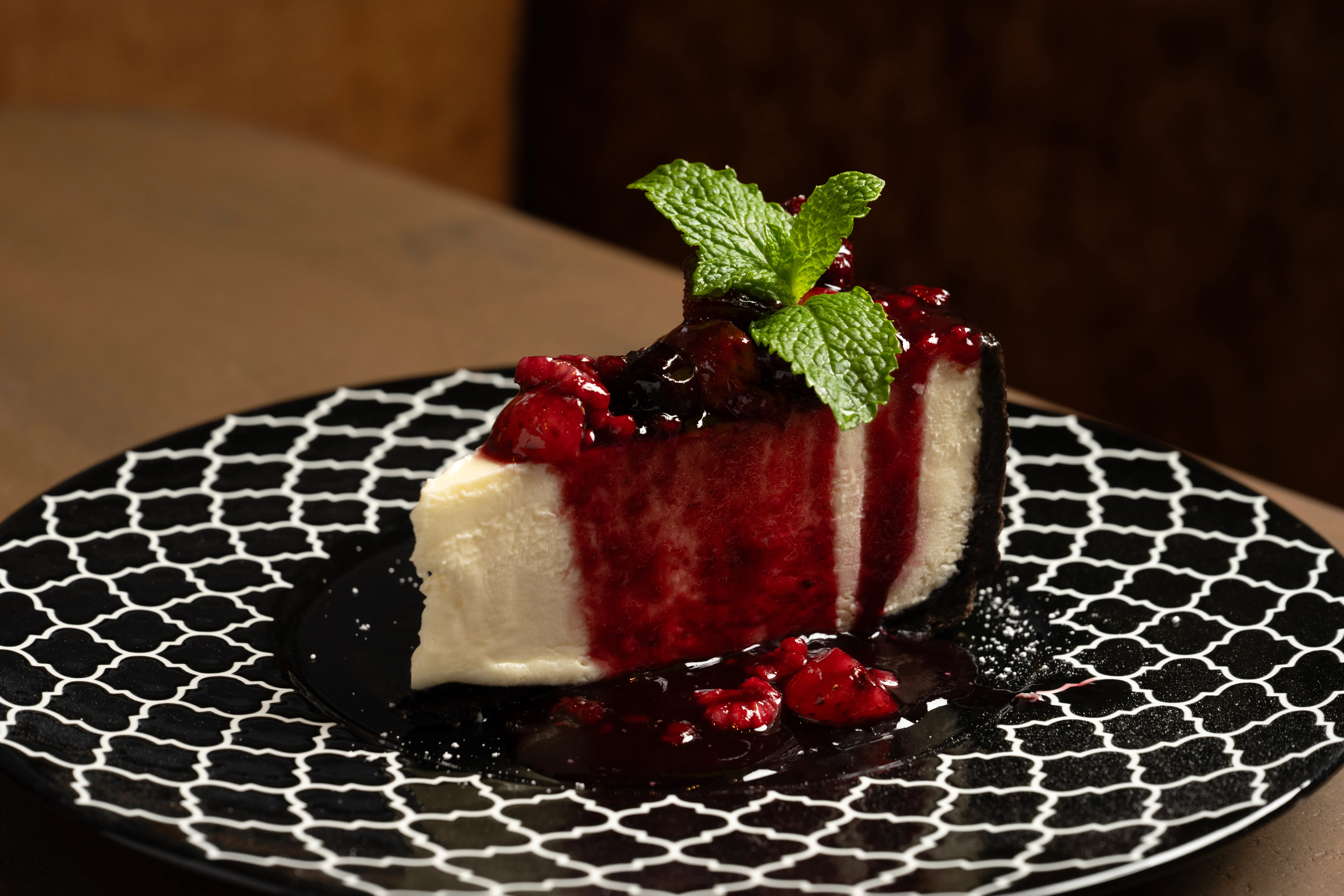 Slice of cheesecake topped with red berry sauce and fresh mint leaves on a black and white patterned plate.
