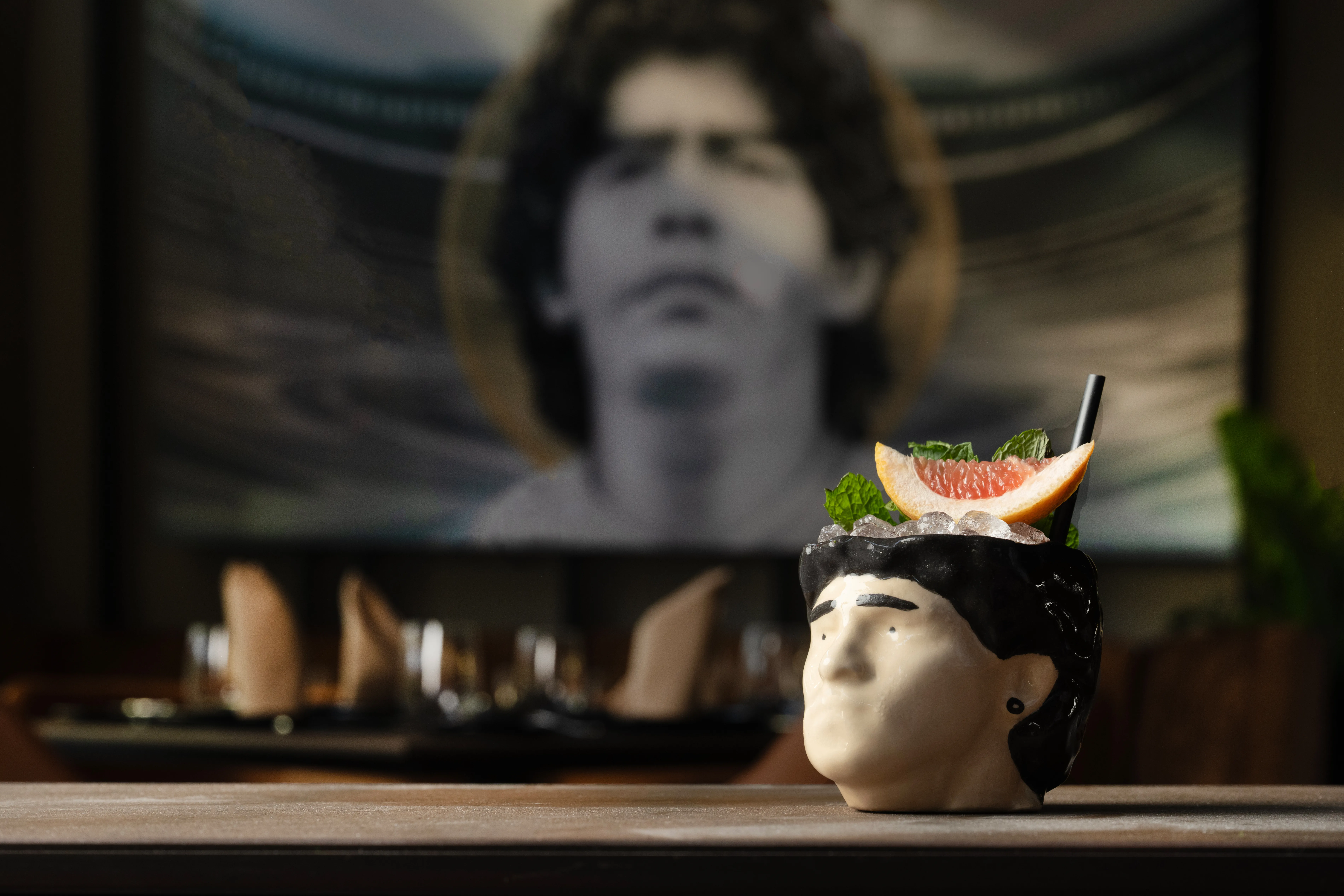 Cocktail served in a ceramic mug shaped like a man's head, garnished with mint and a grapefruit slice, with a blurred portrait of the same man in the background.
