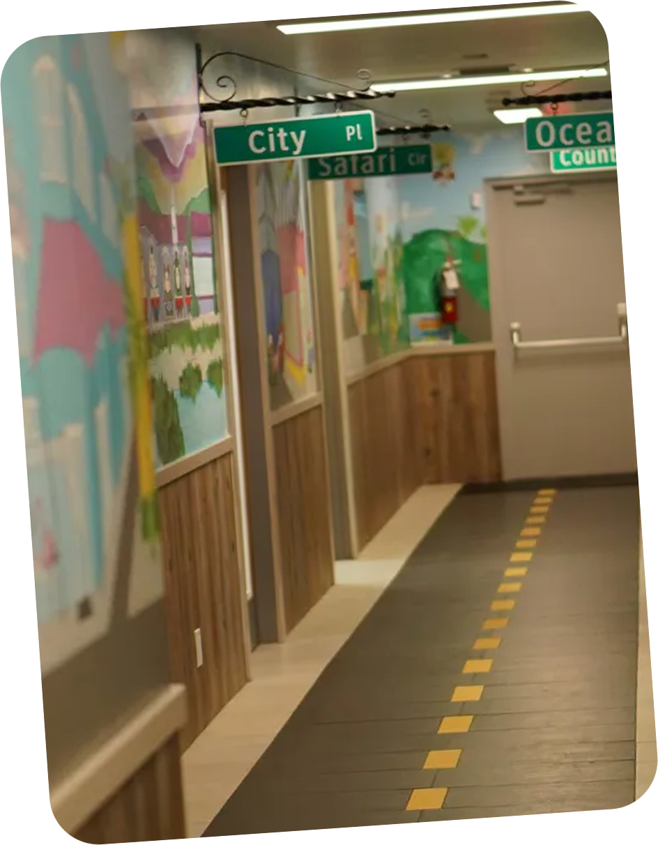 Indoor hallway with murals on walls and overhead green street signs labeled City Pl, Ocean Dr, Safari Cir, and Country Ln, with a yellow dashed line on the floor.