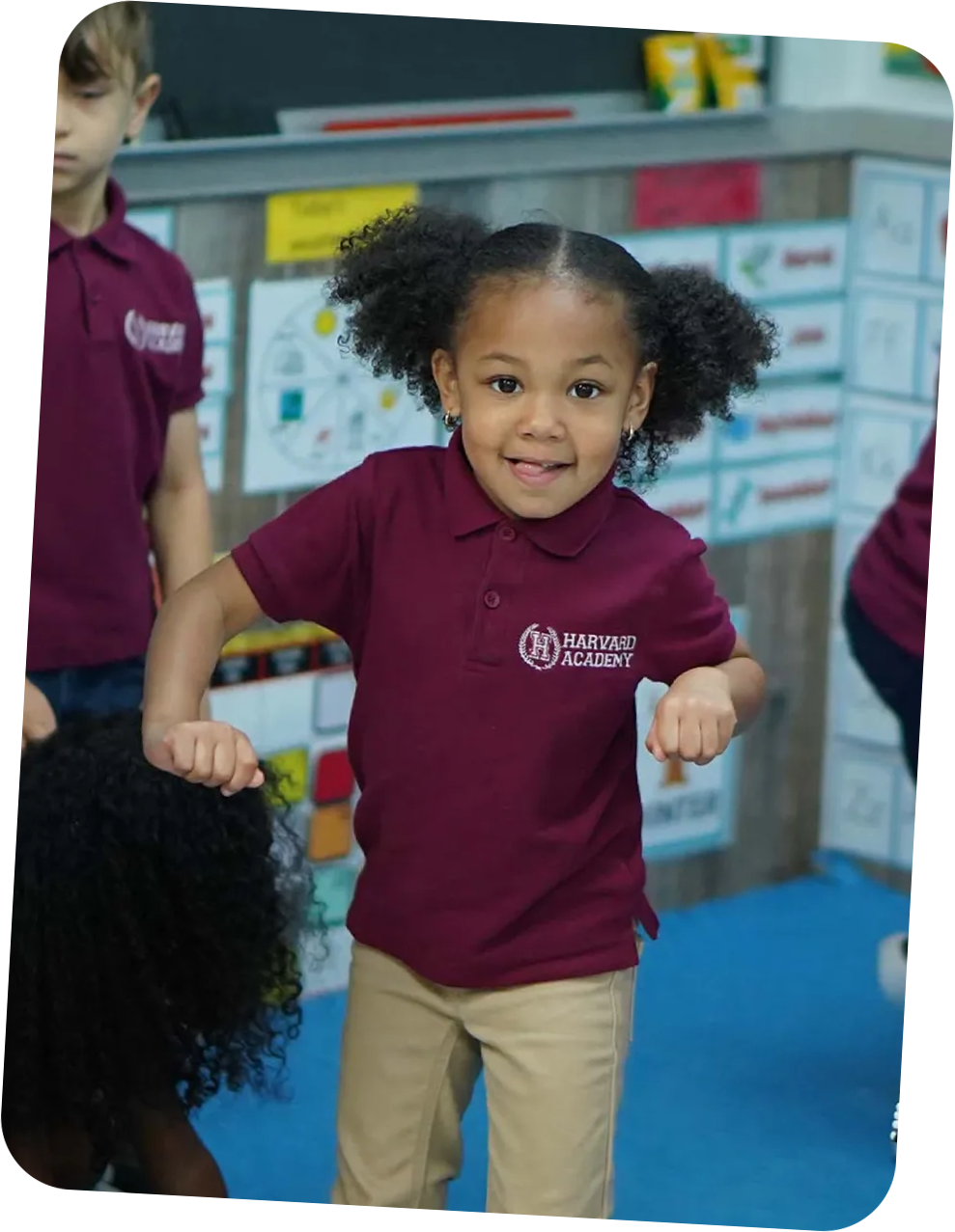 Young girl with curly pigtails smiling and posing in a maroon Harvard Academy polo shirt and beige pants indoors.