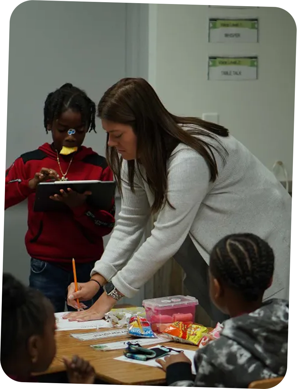 Teacher leaning over a table helping students with schoolwork while one student holds a tablet and another looks on.