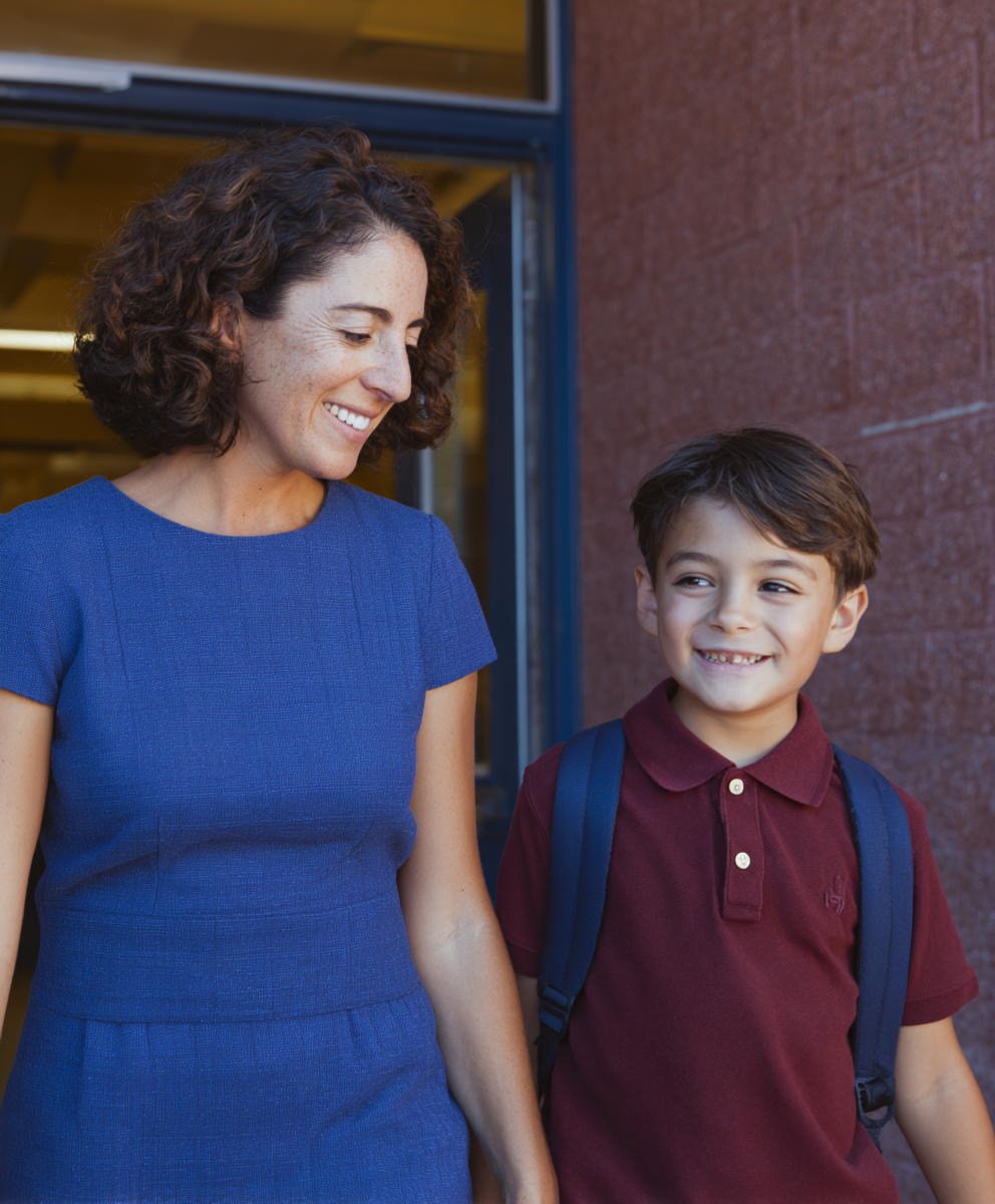 Smiling woman in blue dress holding hands with a young boy wearing a maroon polo and backpack, walking outside near a building.
