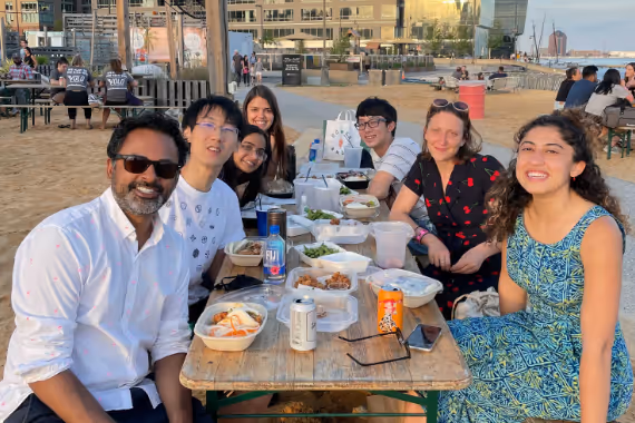A diverse group of seven young adults smiling and seated at an outdoor picnic table with food and drinks in an urban setting.