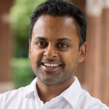 Smiling man with short curly hair and light beard wearing a white shirt, standing outdoors with a blurred natural background.