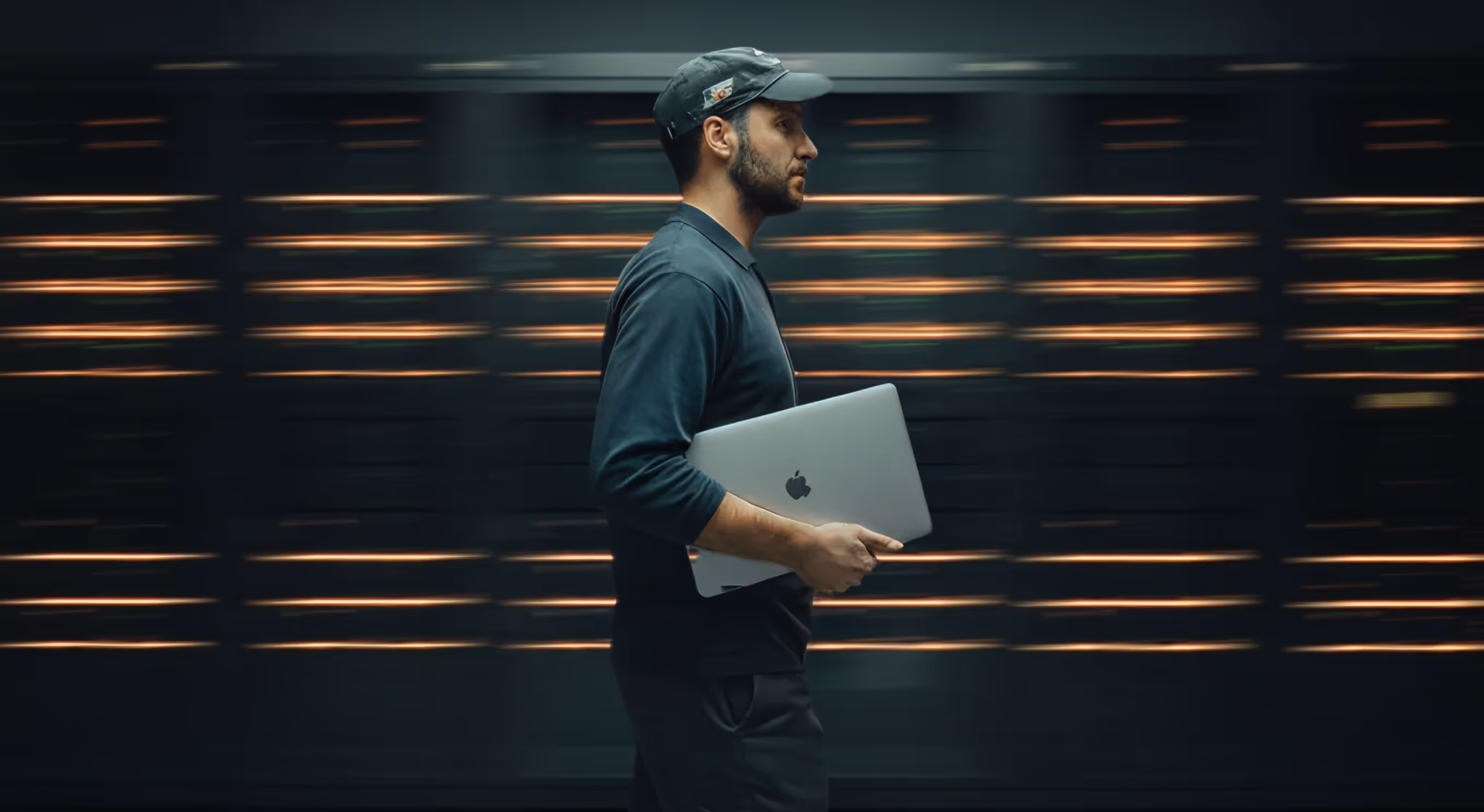 Man wearing a dark cap and shirt holding a closed laptop while walking past a dark background with horizontal orange light lines.