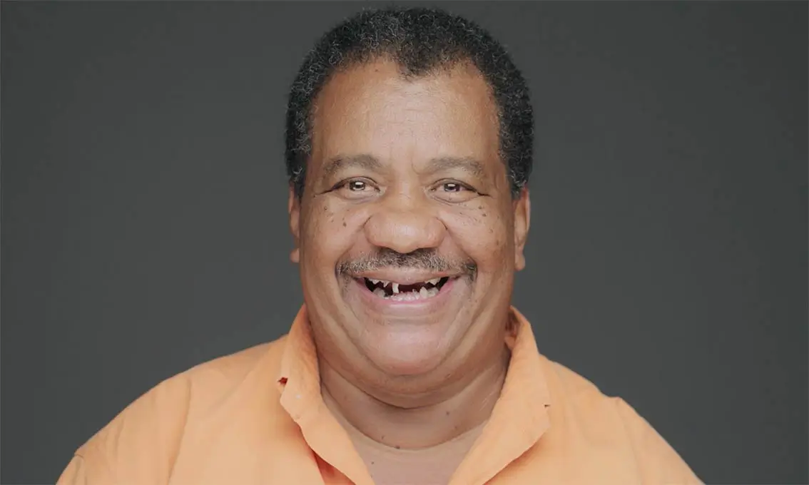 A smiling man with short hair wearing an orange shirt against a plain background.