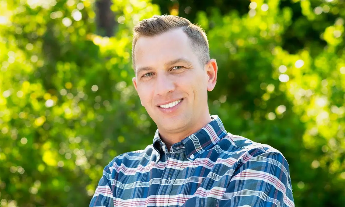 A person smiling outdoors, wearing a plaid shirt with greenery in the background.