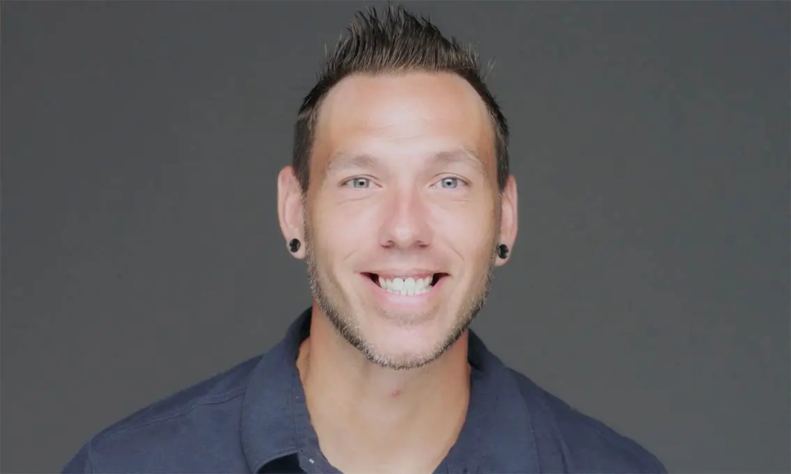 A man with short hair and a navy shirt smiles against a plain gray background.