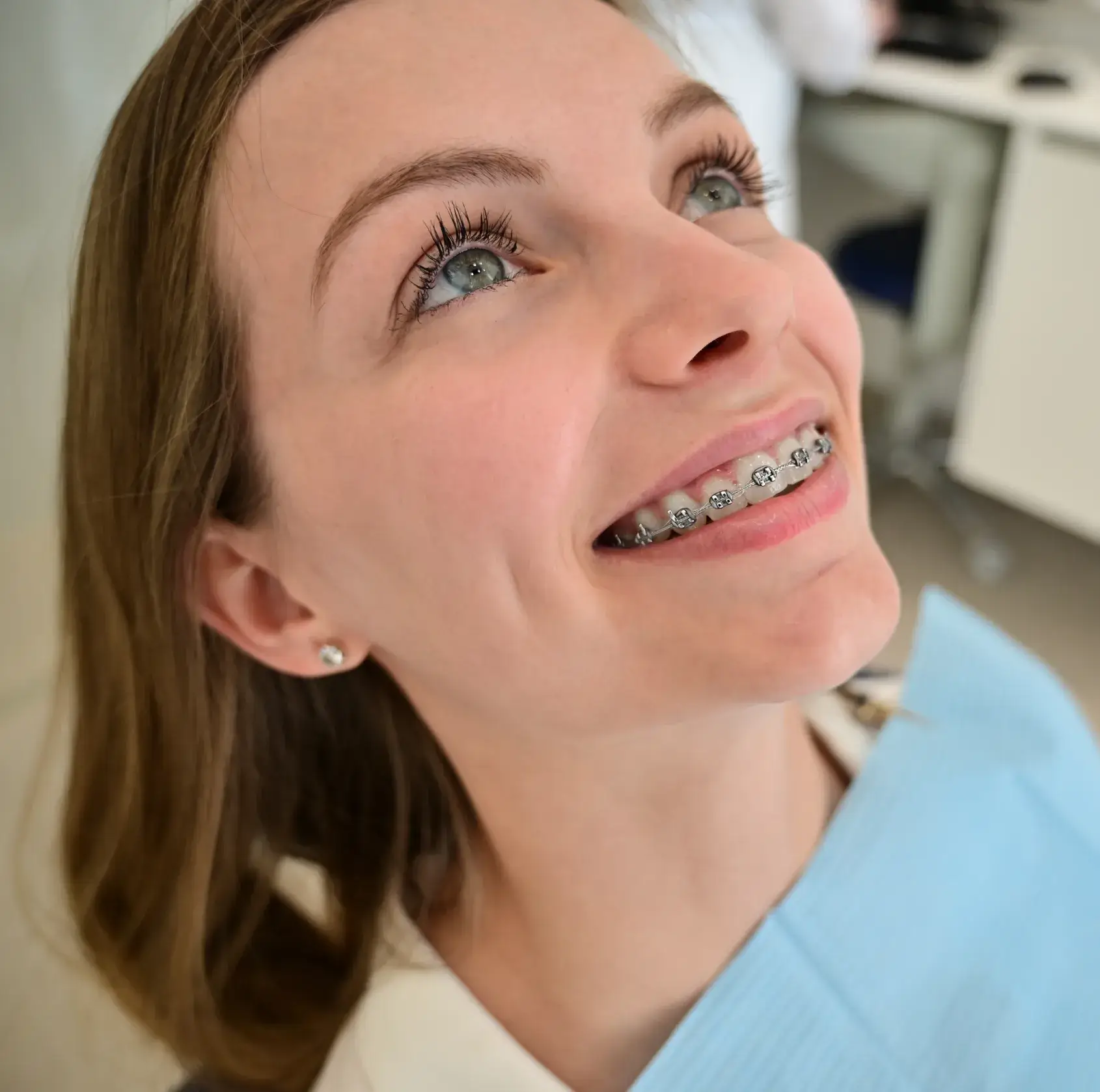 A smiling person with braces looks upward, wearing a dental bib in a clinic setting.
