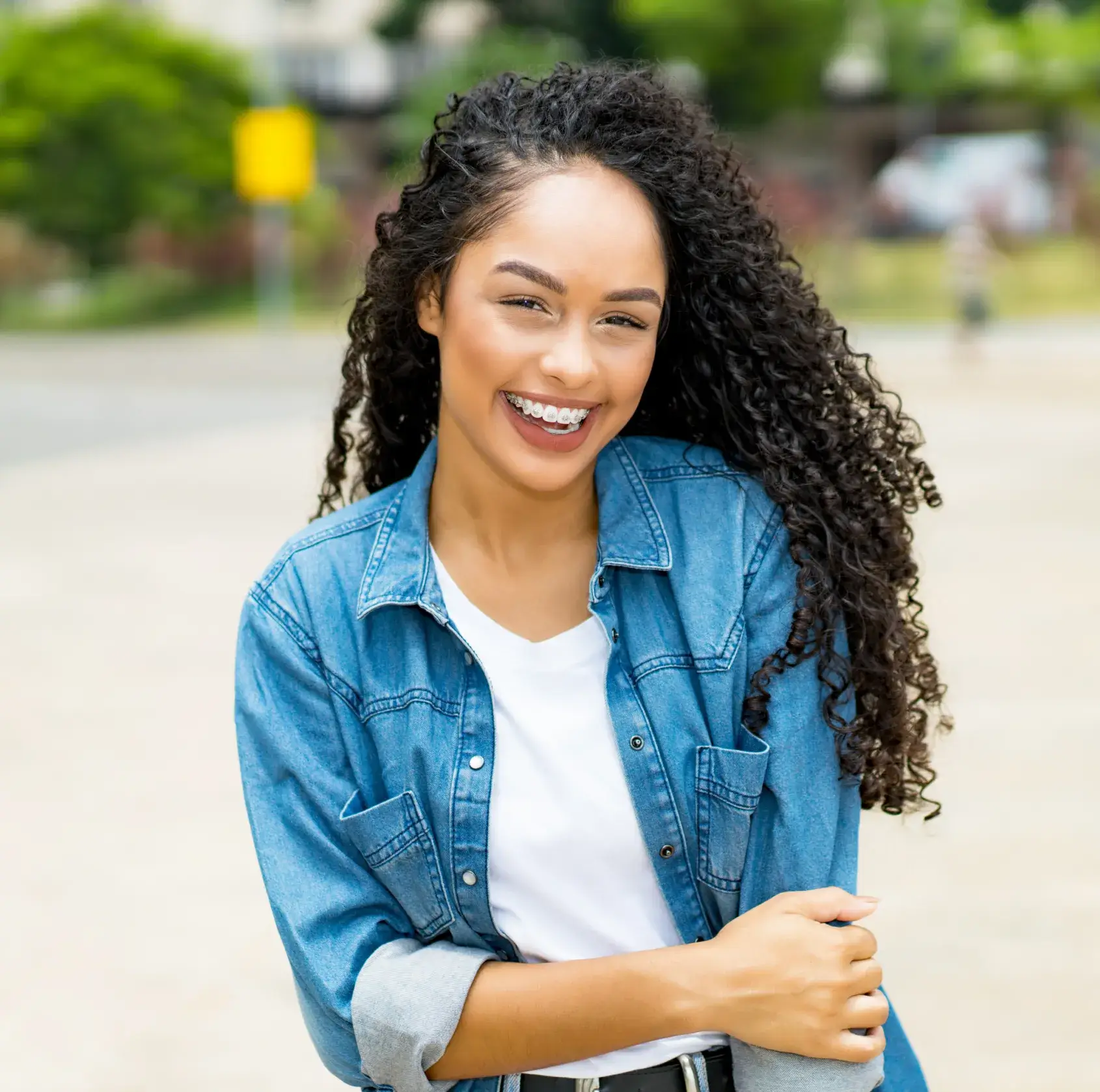 A woman with curly hair smiles while standing outdoors, wearing a denim shirt over a white top.
