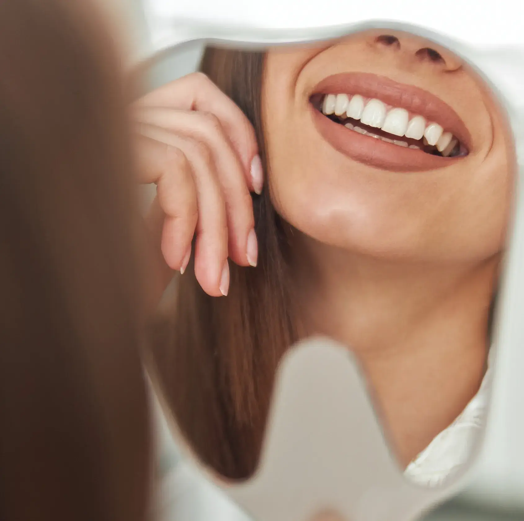 A woman smiles brightly, reflected in a handheld mirror.