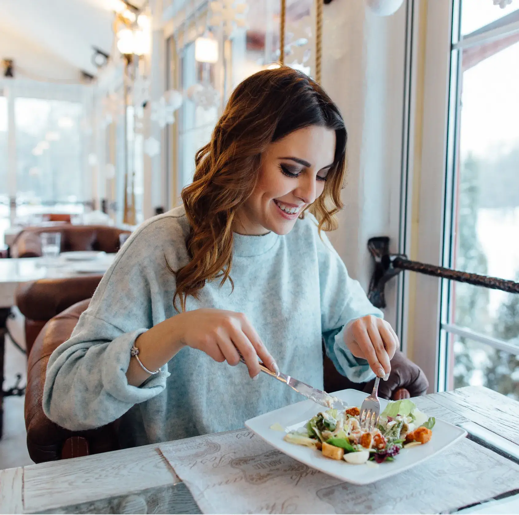 A woman in a restaurant is smiling while eating a salad from a white plate.