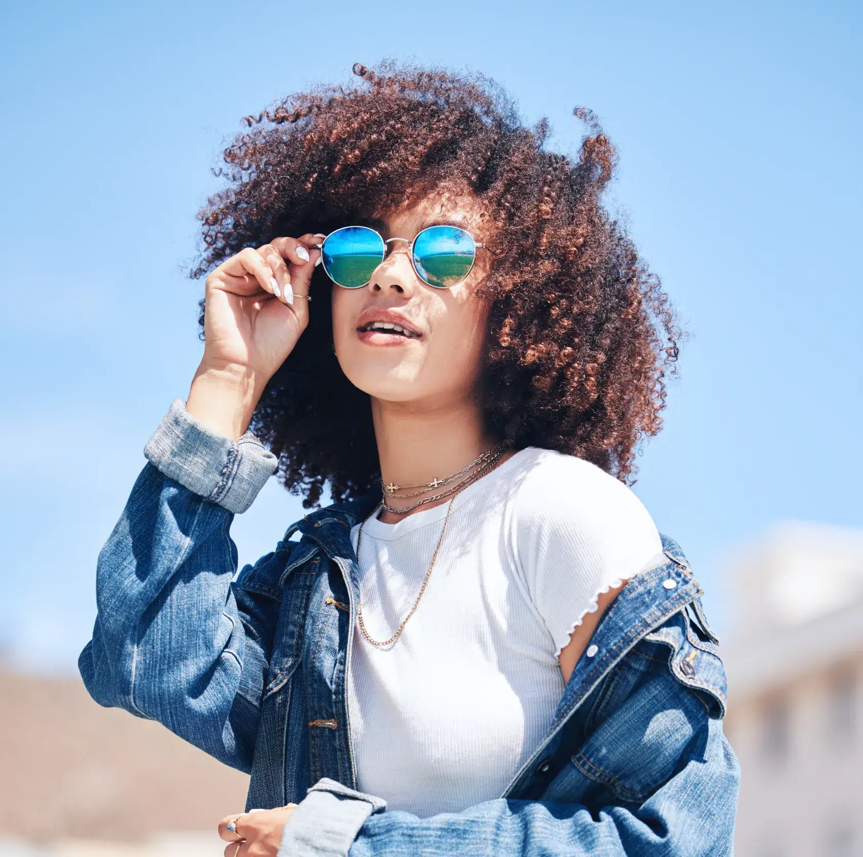 Young woman with curly hair adjusts sunglasses while wearing a denim jacket and white shirt.