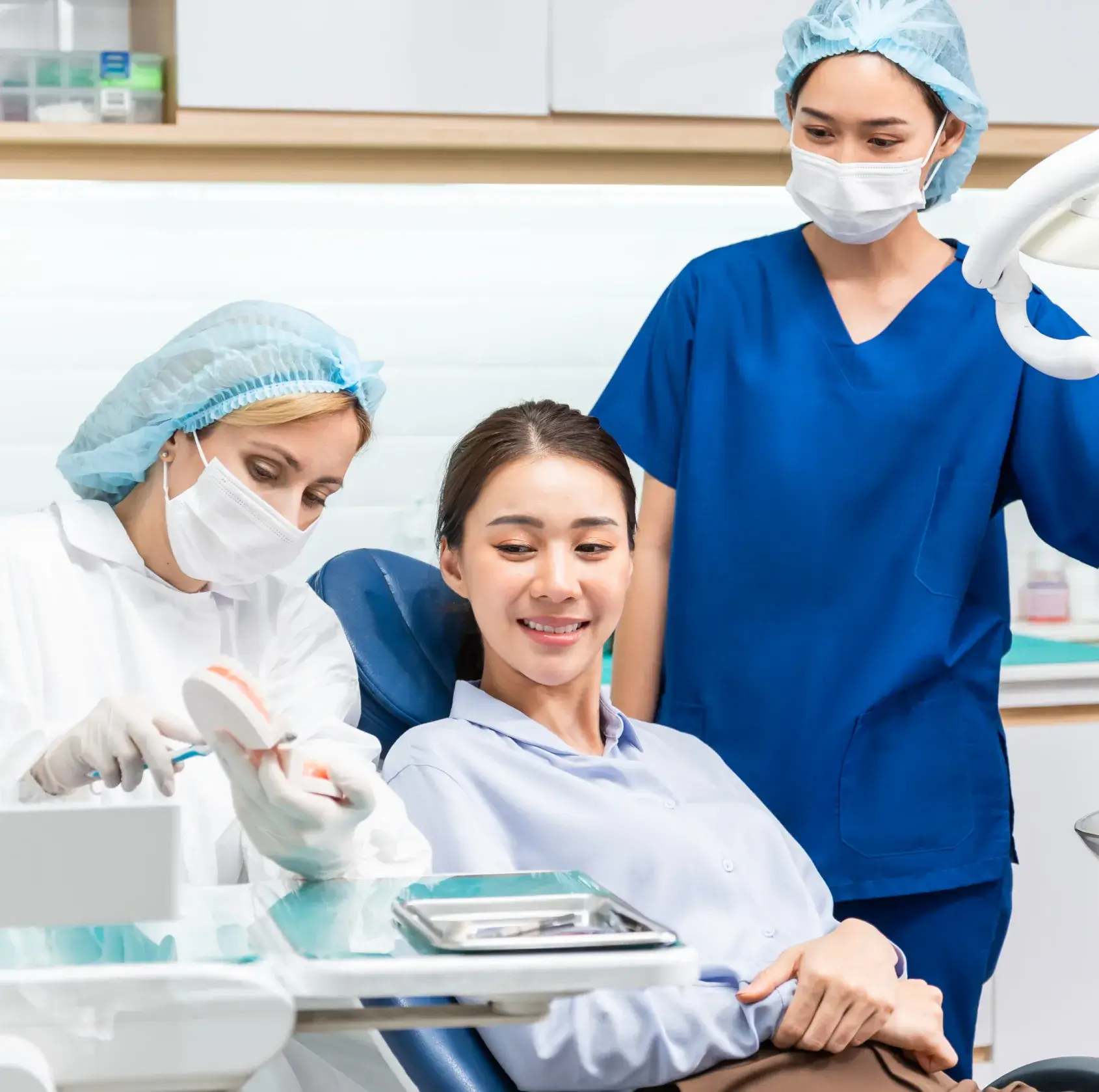 A dentist shows a model of teeth to a patient, with a nurse observing nearby.