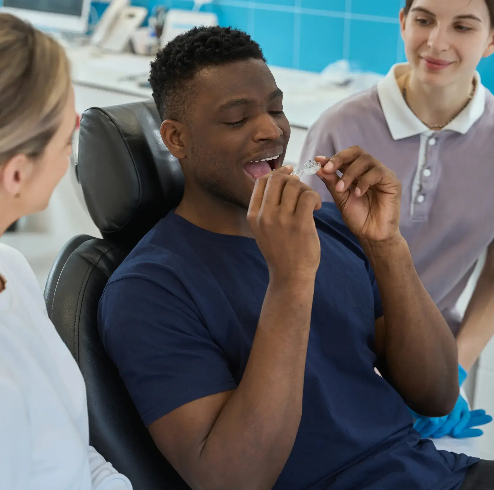 A man in a dental chair holds an orthodontic aligner, assisted by two dental professionals.
