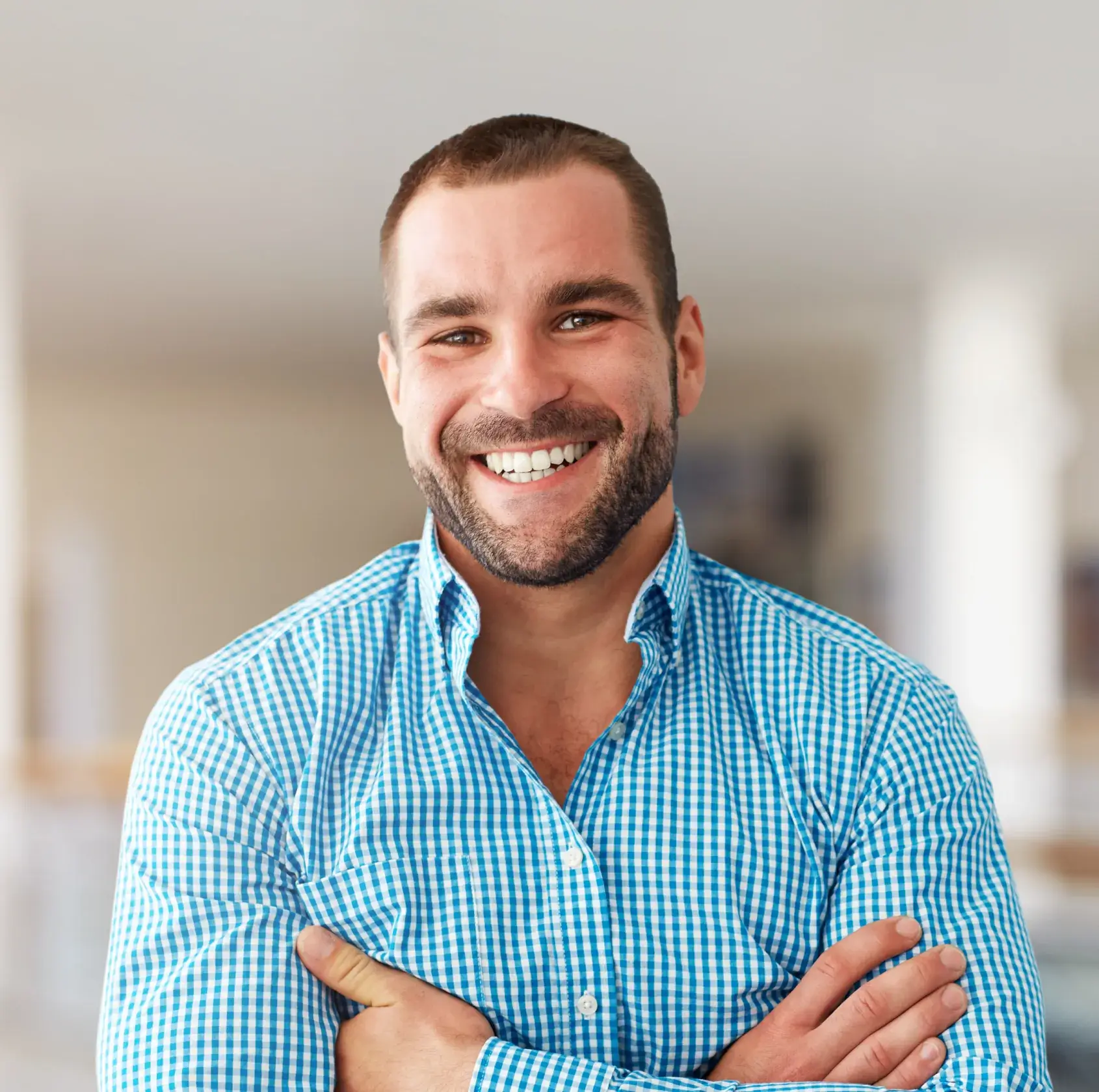 A man in a blue checkered shirt smiles with arms crossed.