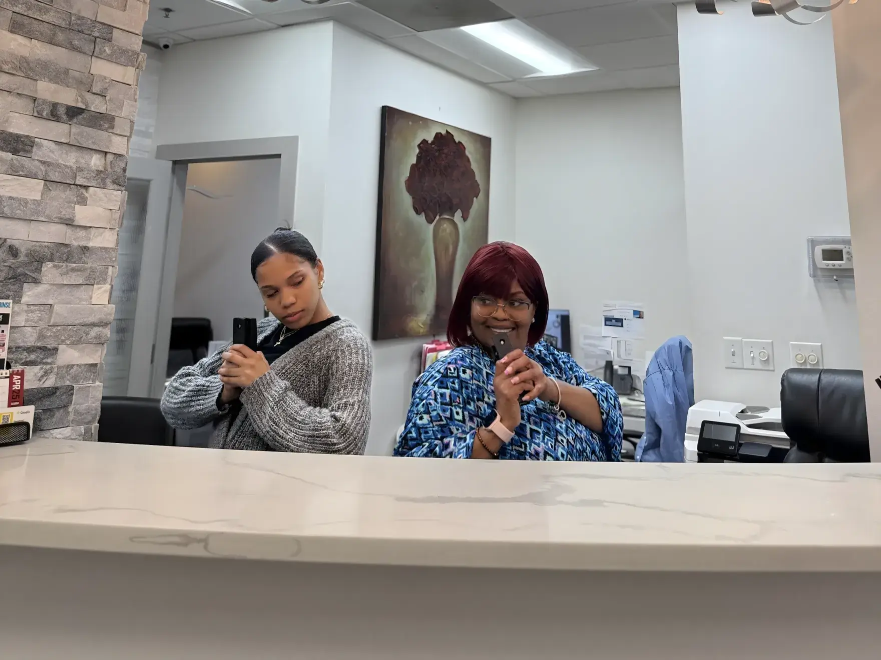 Two people standing behind a counter, both looking into a mirror and taking selfies.