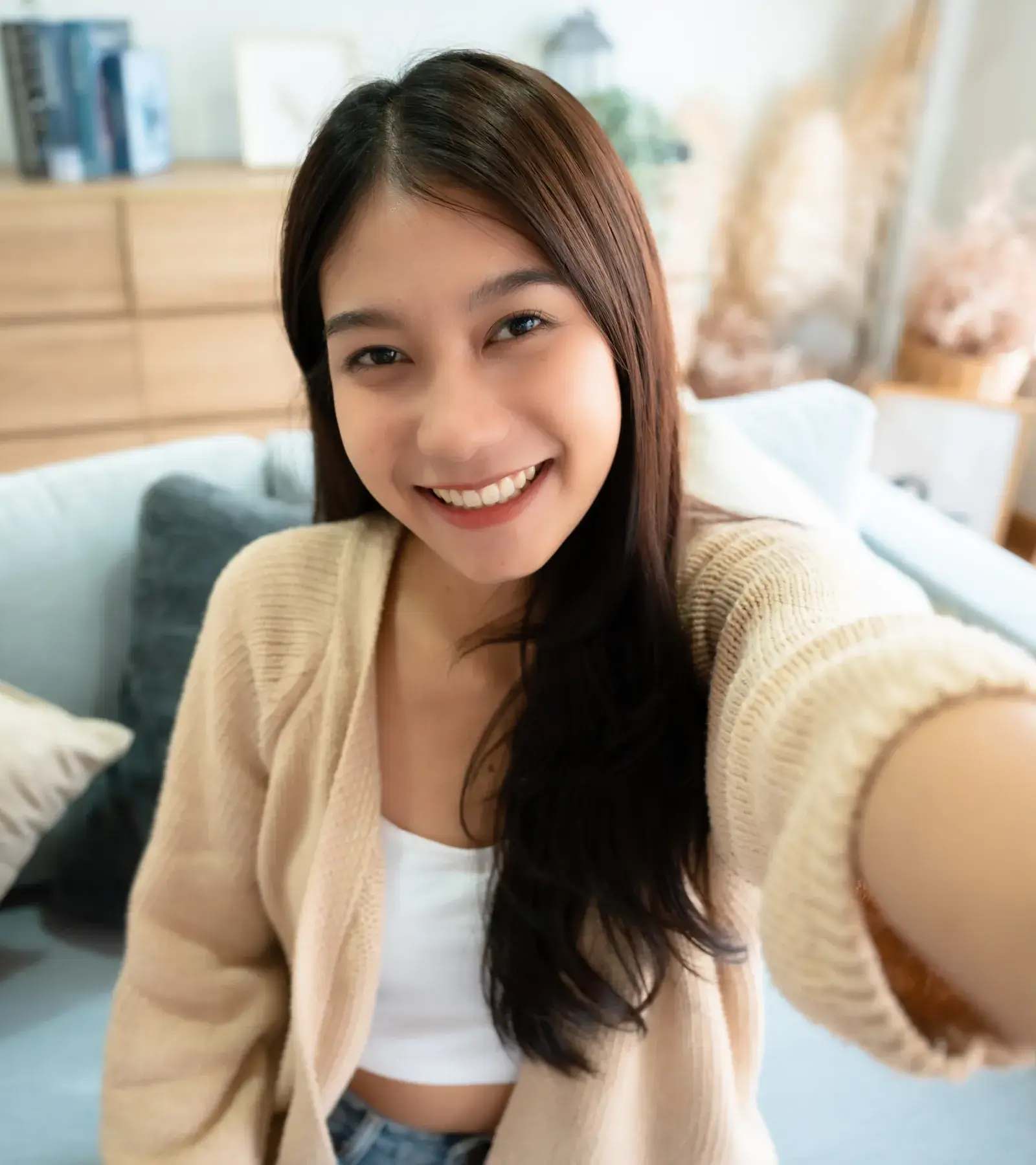 A woman with long hair is smiling while taking a selfie in a cozy room.
