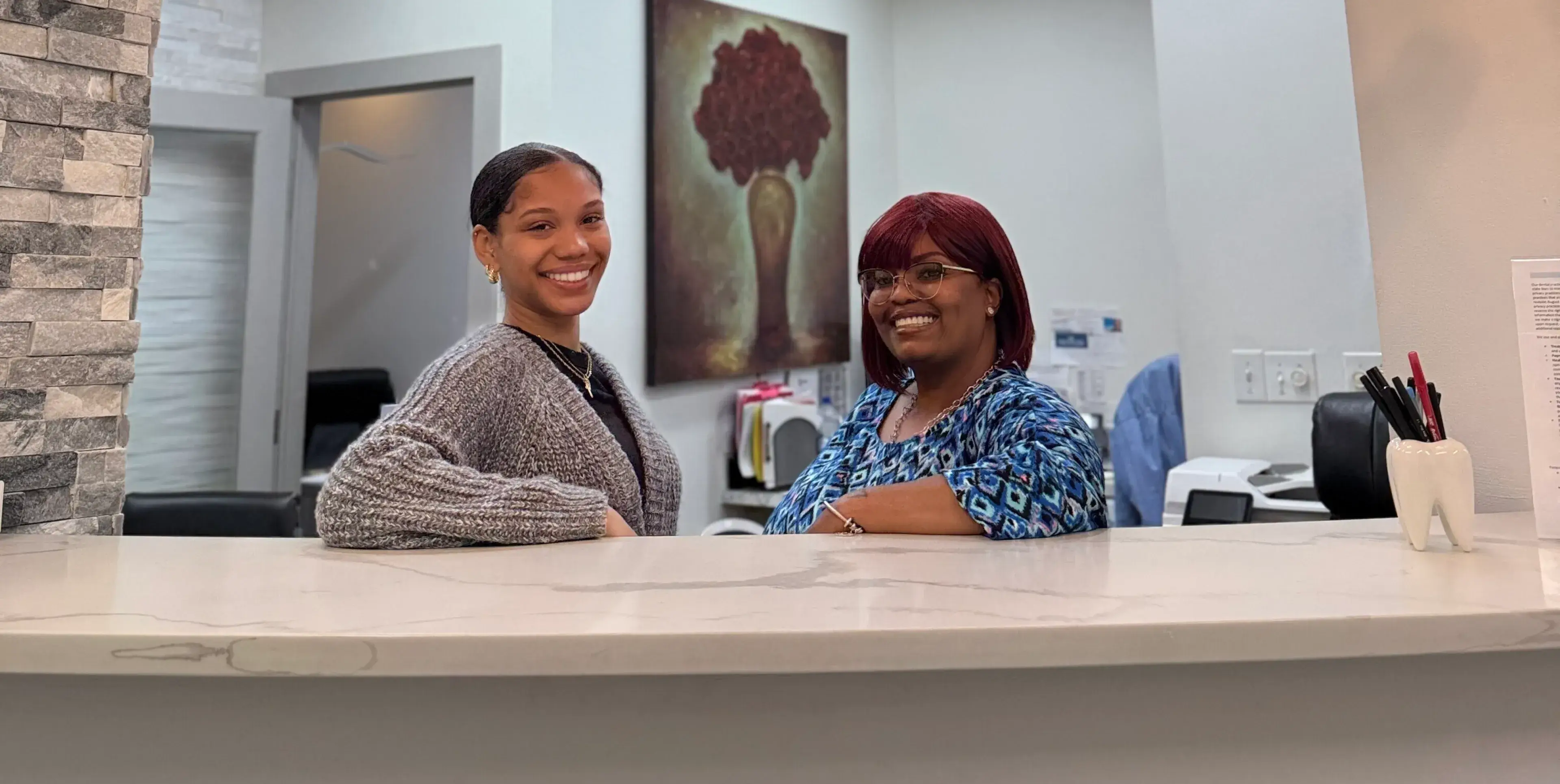 Two women smile while standing behind a reception desk in an office.