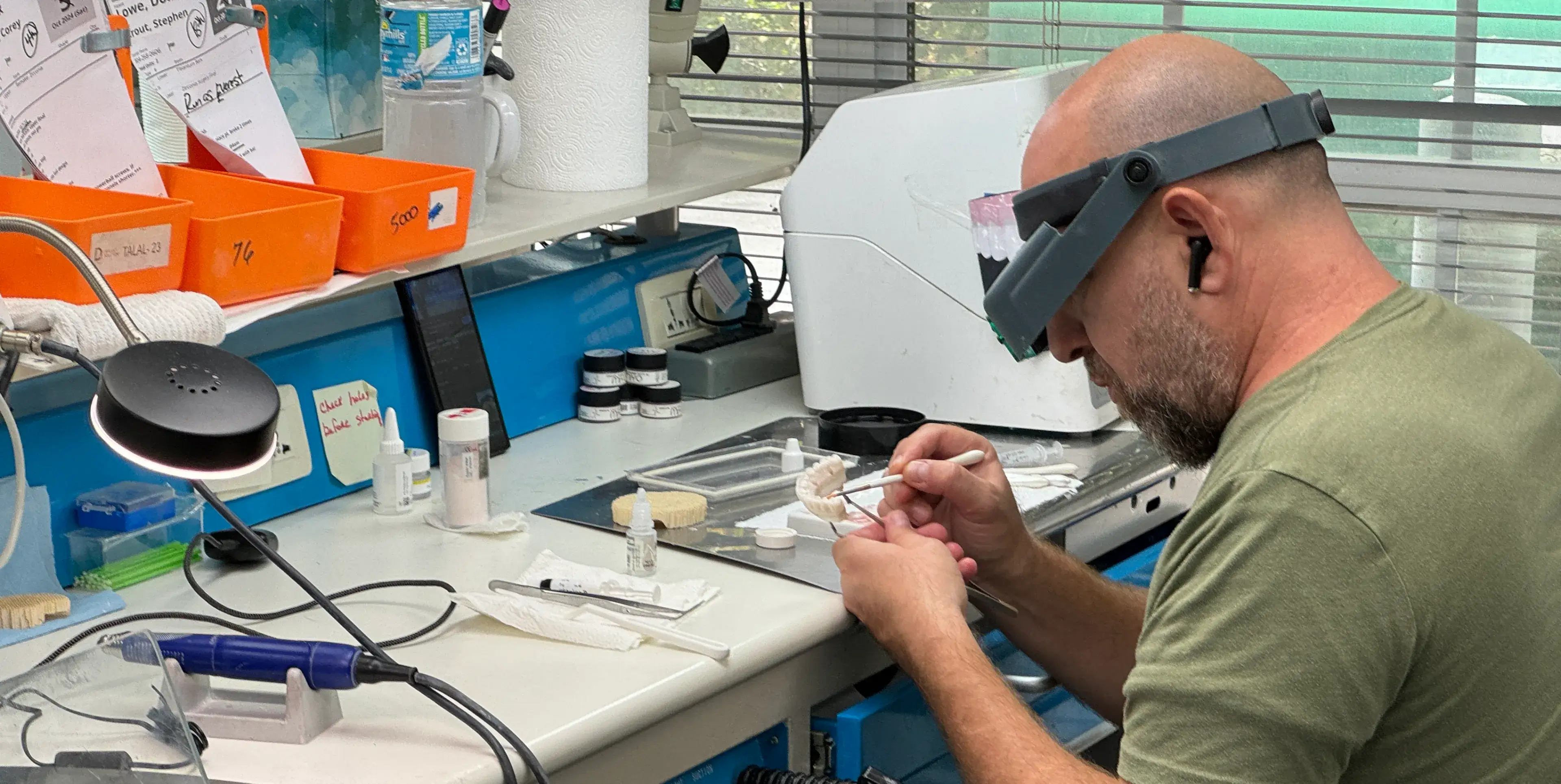 A person wearing magnifying glasses works on a dental model at a lab station.