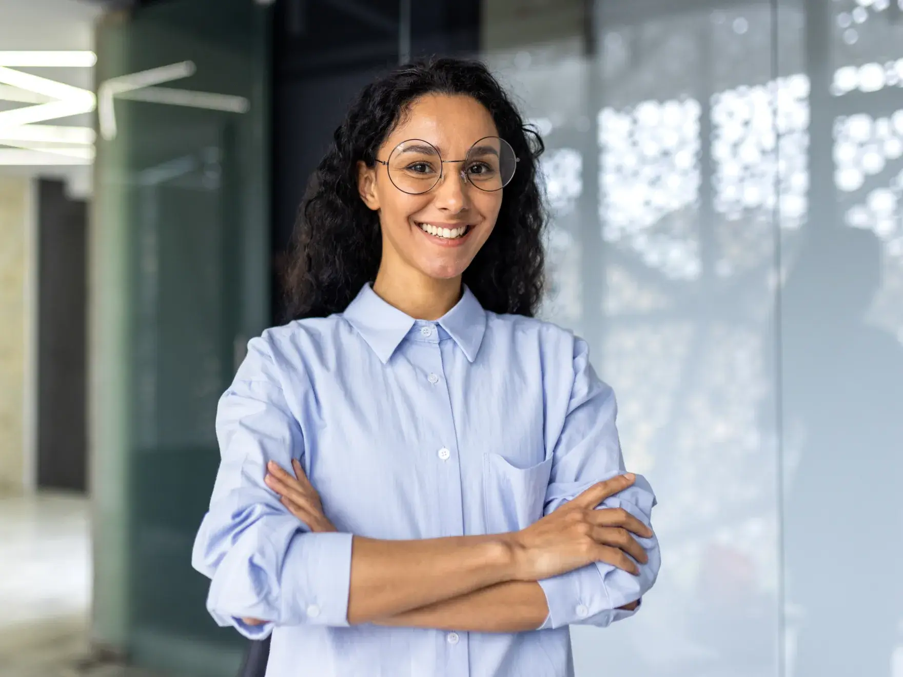A person with glasses smiles confidently, standing with arms crossed in an office setting.