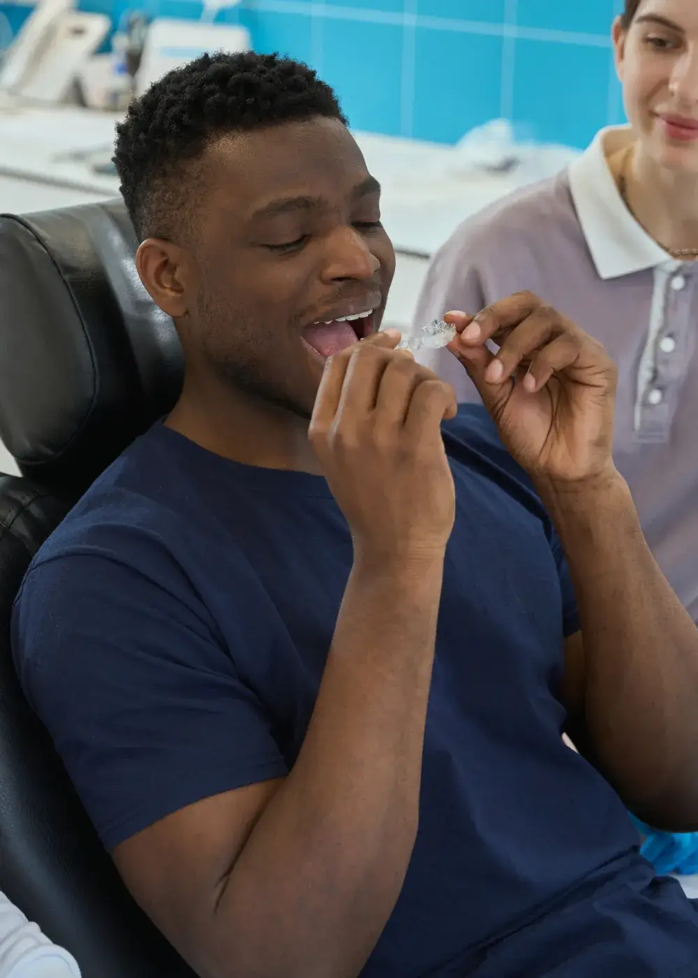 Man in dental chair is about to insert a clear dental aligner while a person observes.