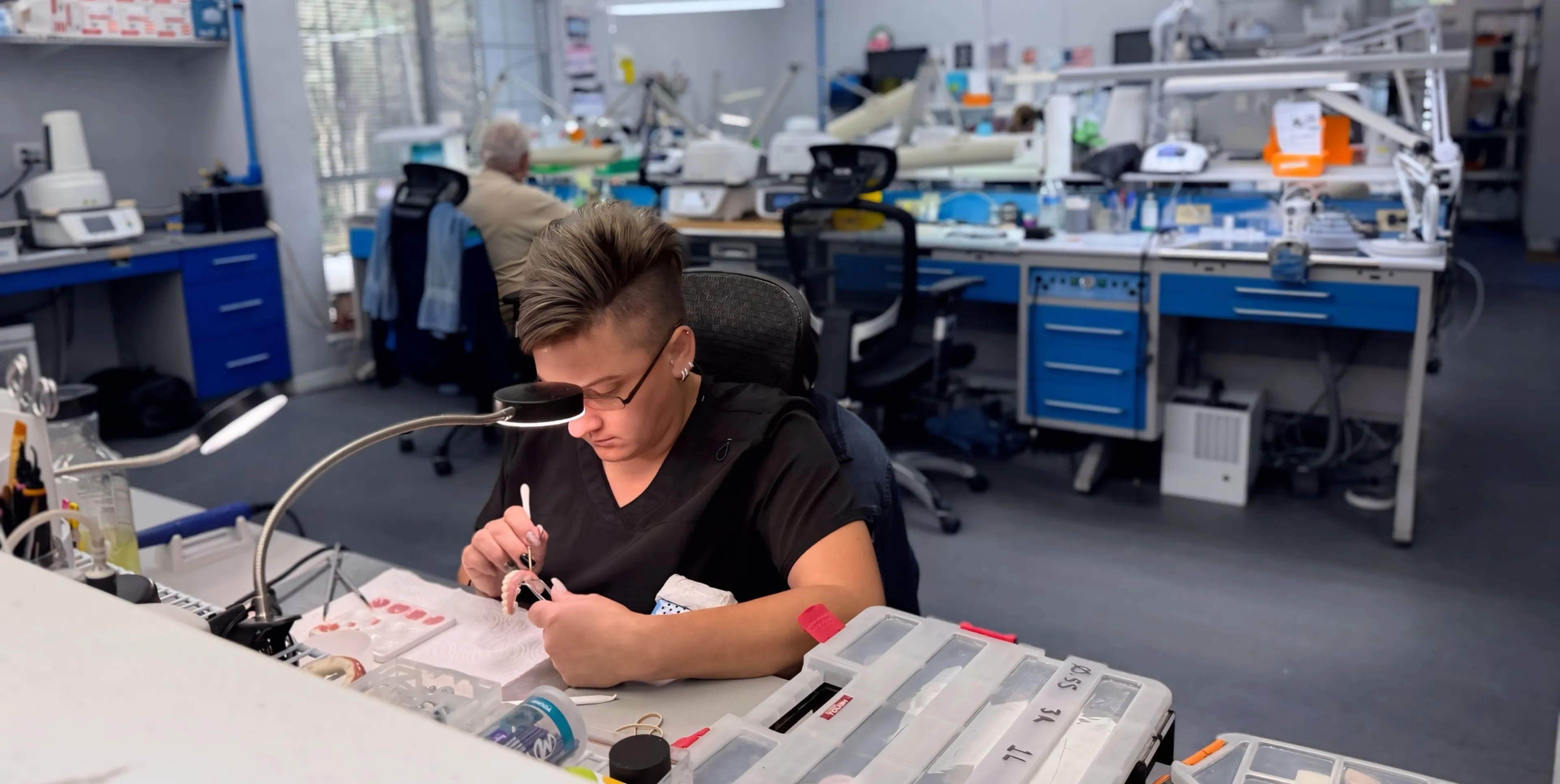 Person focused on detailed work at a desk in a lab with equipment and tools around.