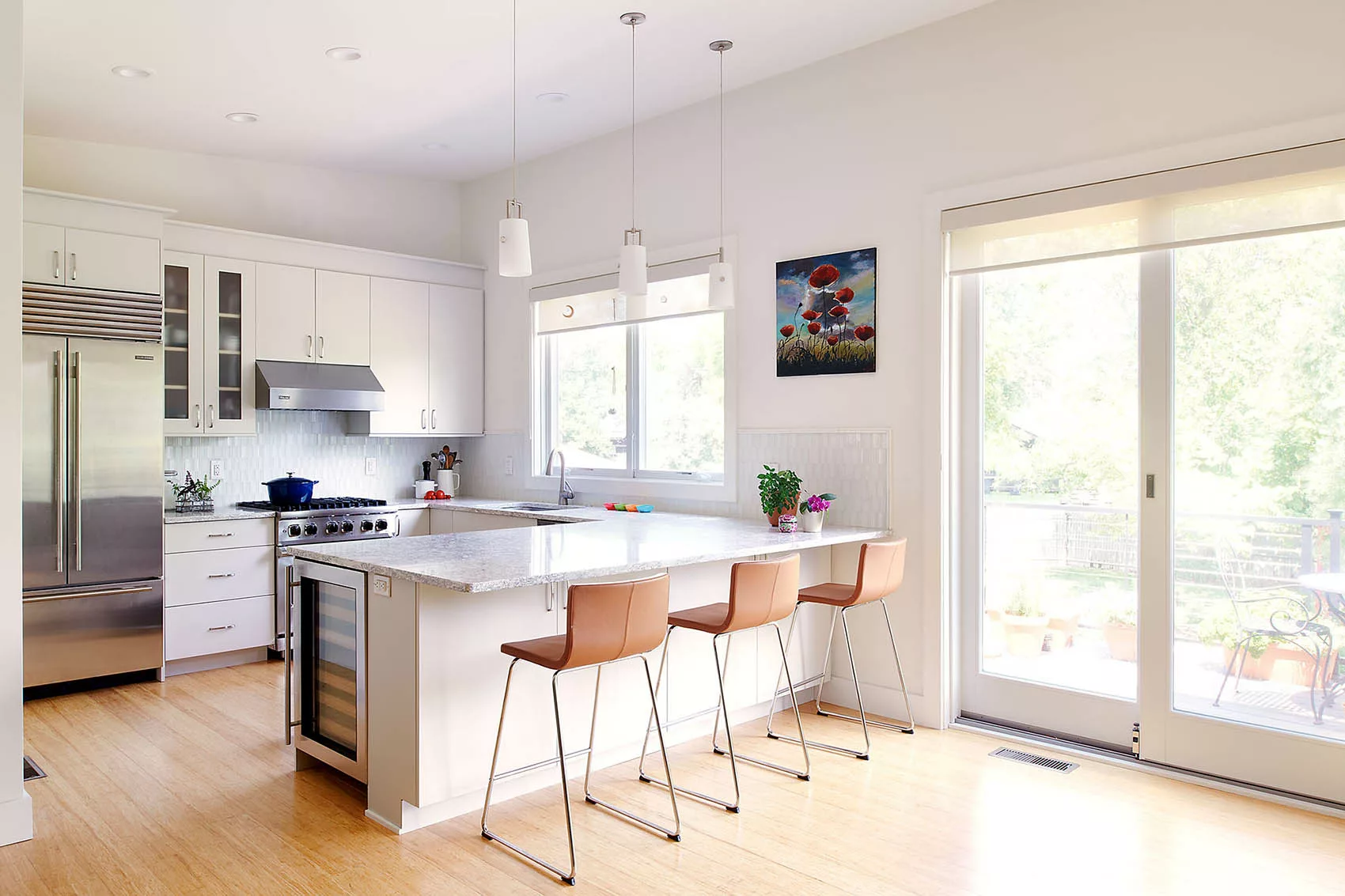 Bright modern kitchen with white cabinets, stainless steel appliances, marble island with three brown bar stools, and large sliding glass door.