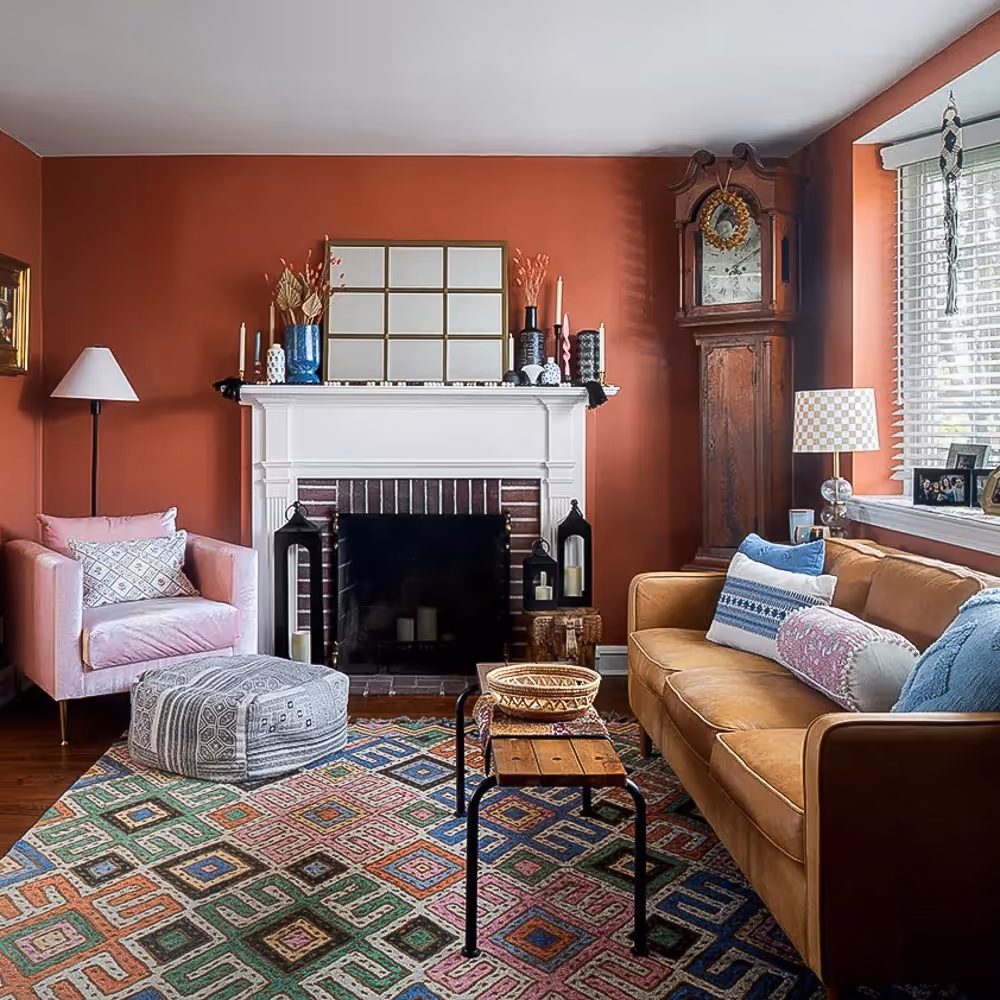 Cozy living room with rust-colored walls, a white fireplace, pink armchair, patterned rug, leather sofa with pillows, and a grandfather clock.