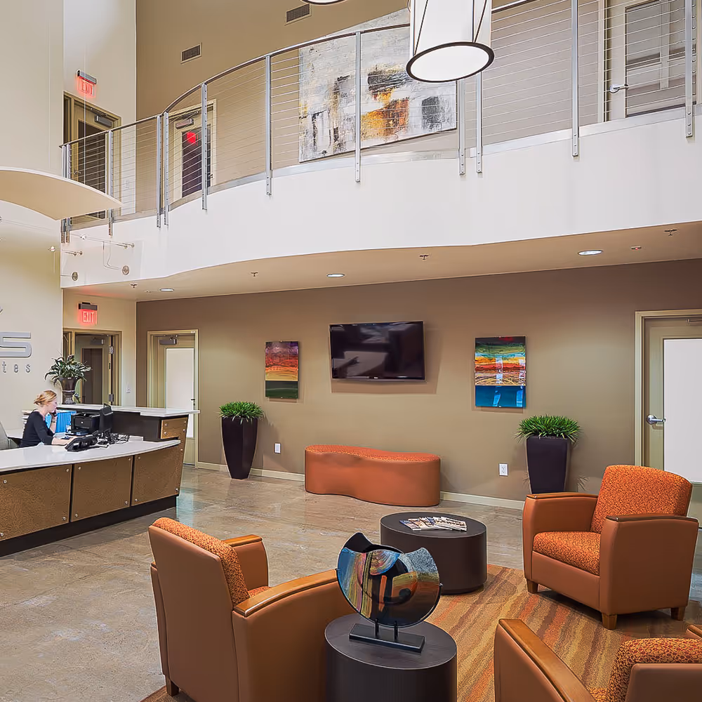 Modern office lobby with a receptionist at the desk, orange armchairs, wall-mounted TV, abstract art, and potted plants.