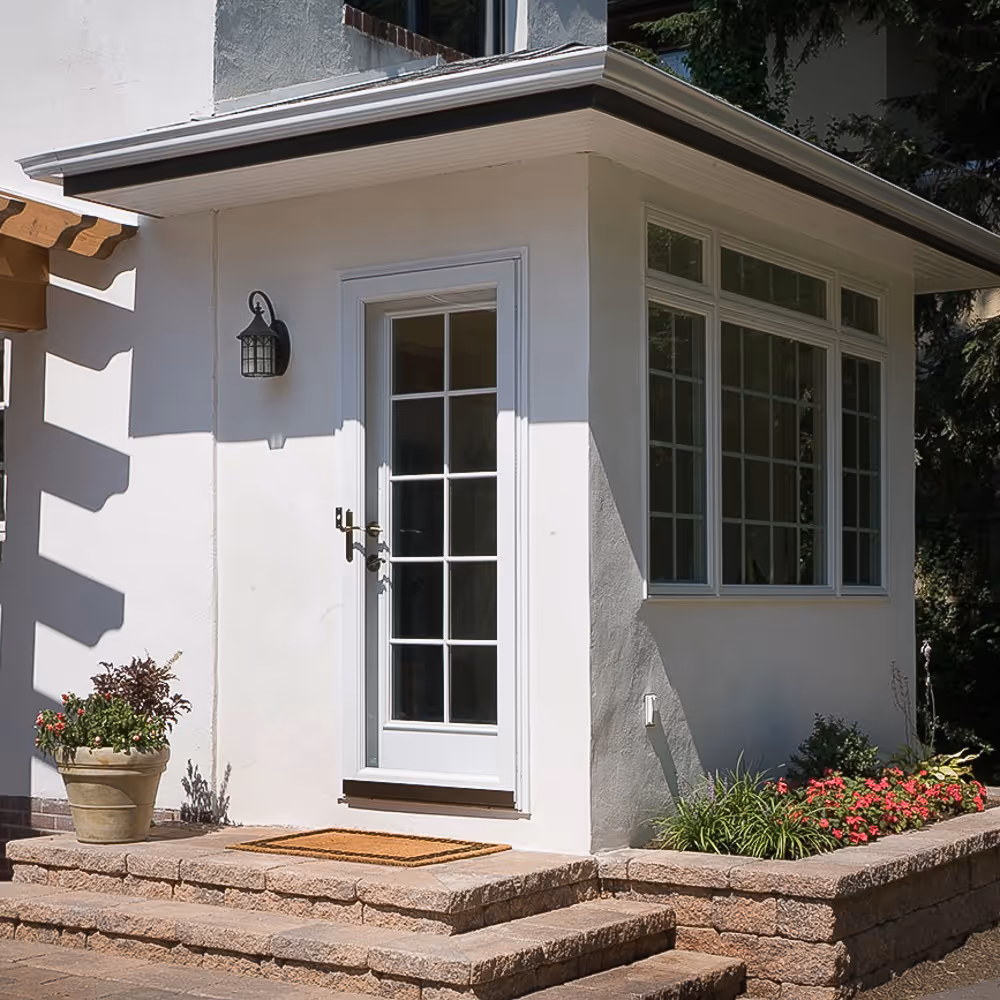 White house entrance with a glass door, stone steps, a doormat, potted plants, and a flower bed.