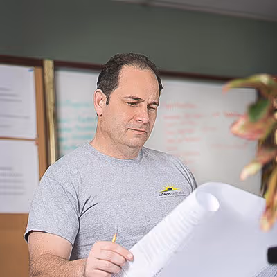 Man in gray t-shirt reviewing a large sheet of paper with notes in a room with a whiteboard.