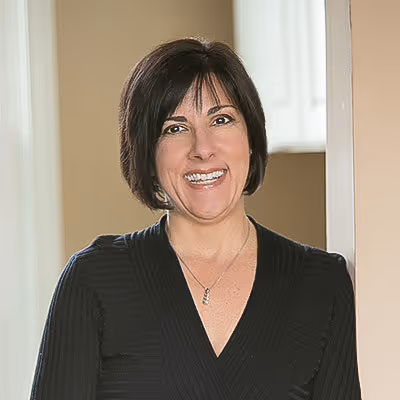 Smiling woman with short dark hair wearing a black top and silver necklace, standing indoors by a doorway.