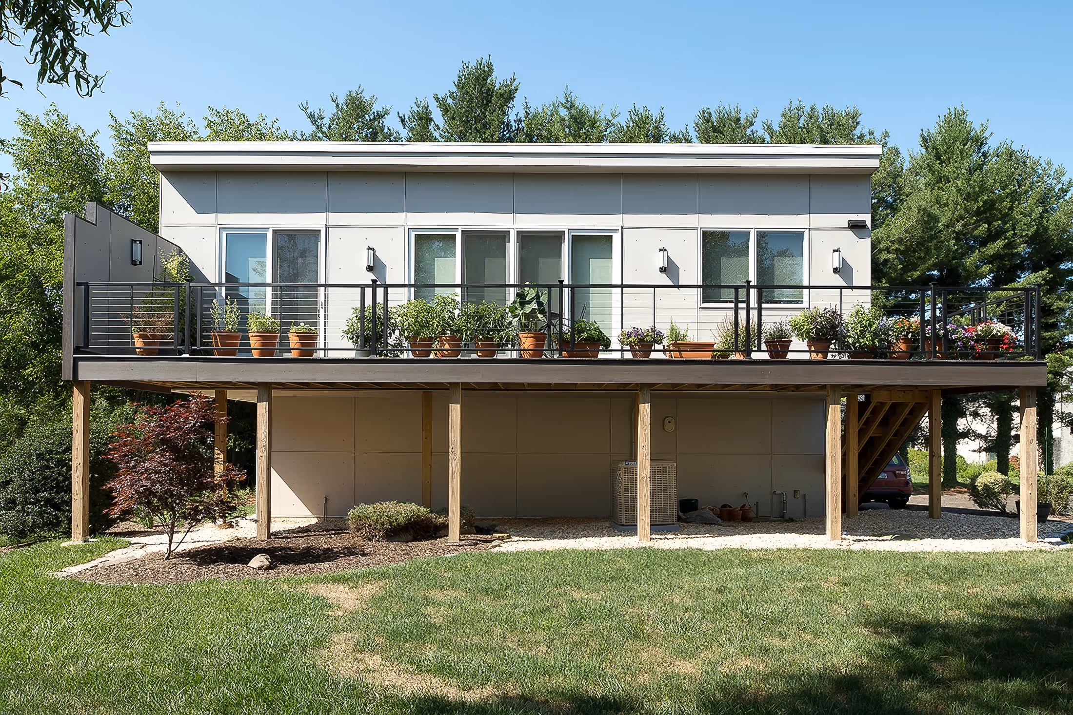 Modern elevated house with a large balcony featuring numerous potted plants, surrounded by grass and trees under a clear blue sky.