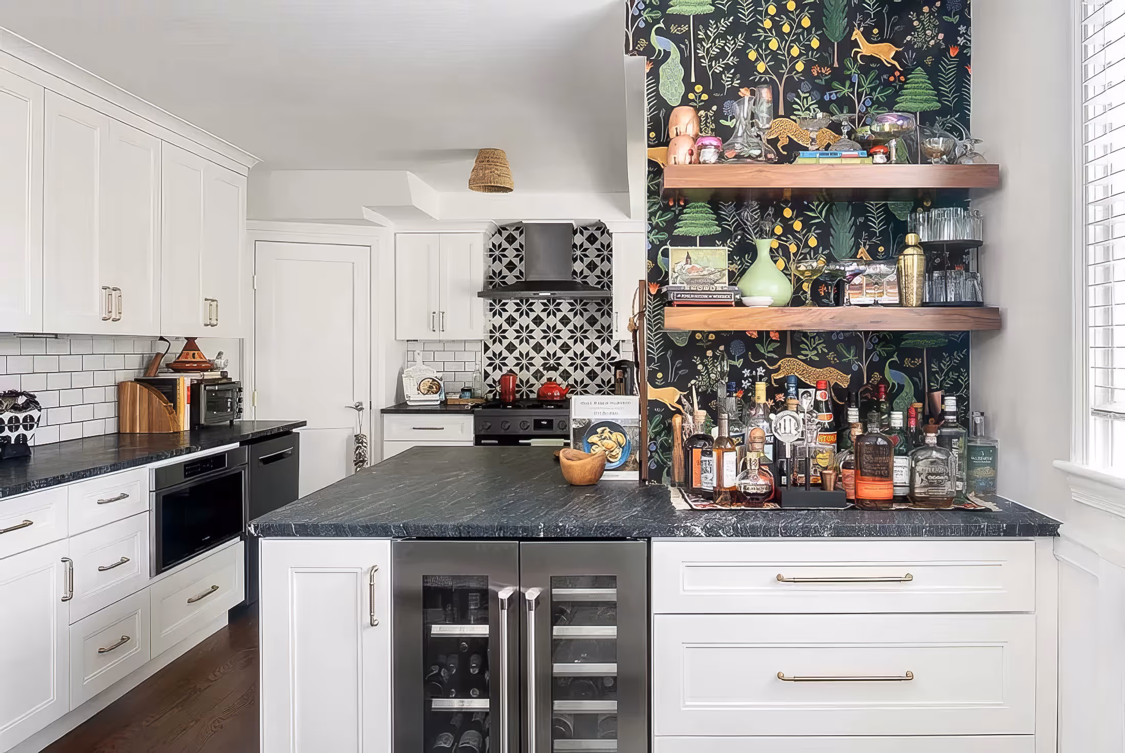 Modern kitchen with white cabinets, black countertops, patterned tile backsplash, and a small bar area with bottles and wooden shelves against a dark floral wallpaper.