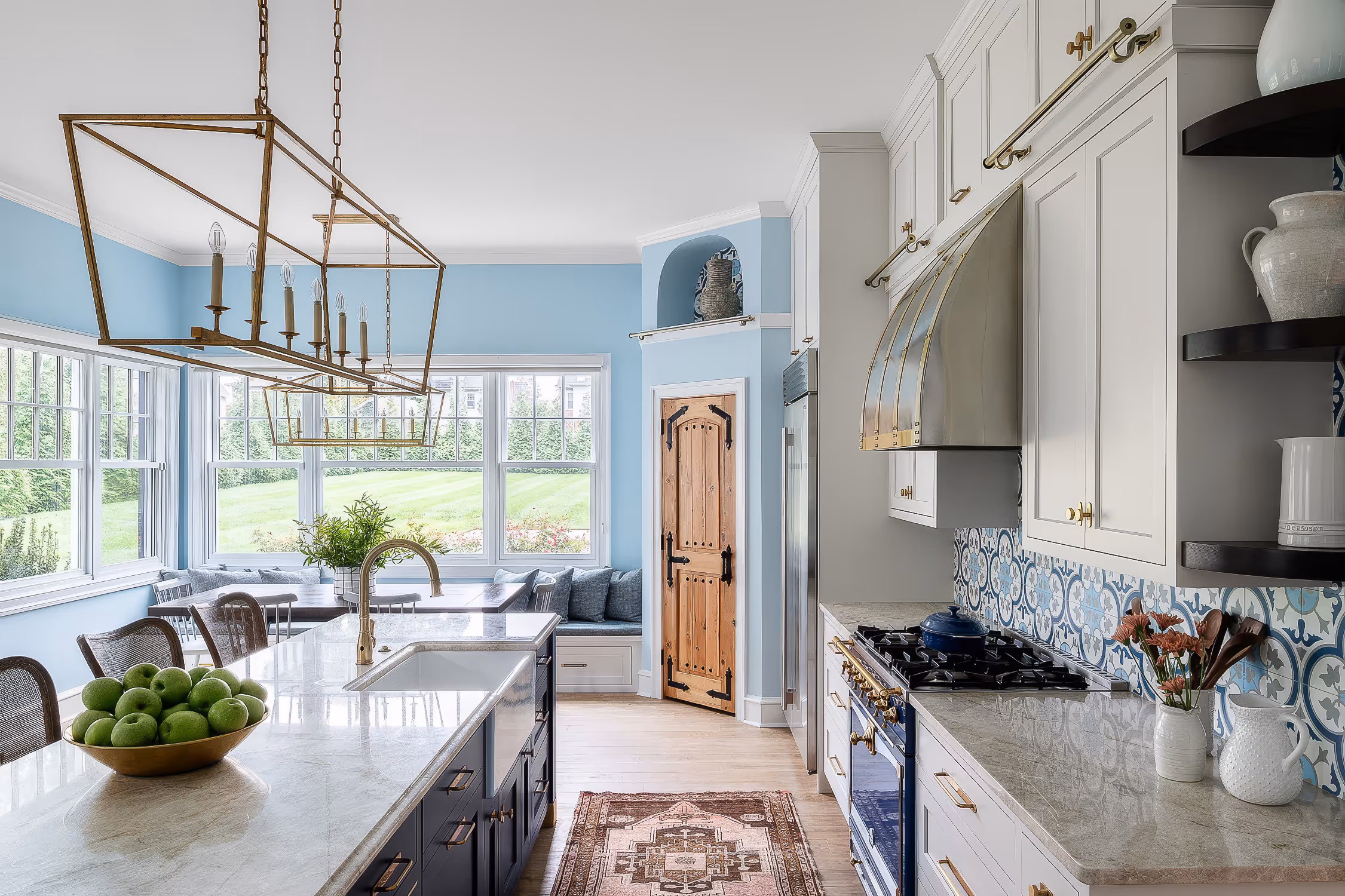 Bright modern kitchen with light blue walls, white and navy cabinets, a marble island with a bowl of green apples, and large windows overlooking a green lawn.