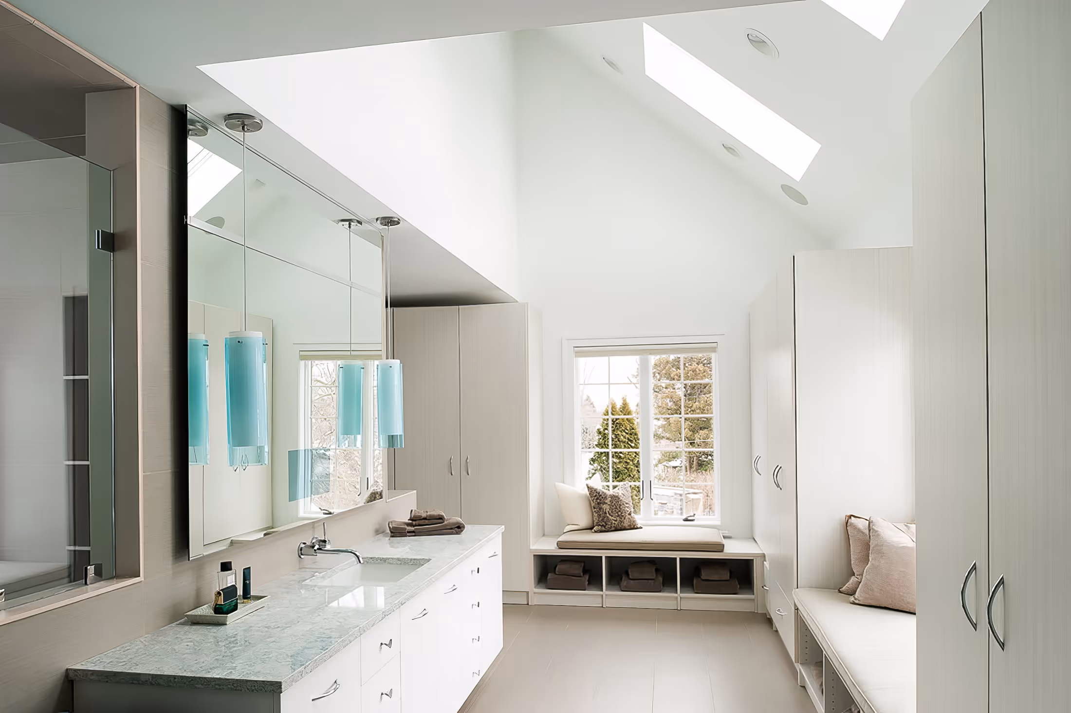 Bright modern bathroom with skylights, large mirror, white cabinets, dual seating benches, and a window seat with cushions.