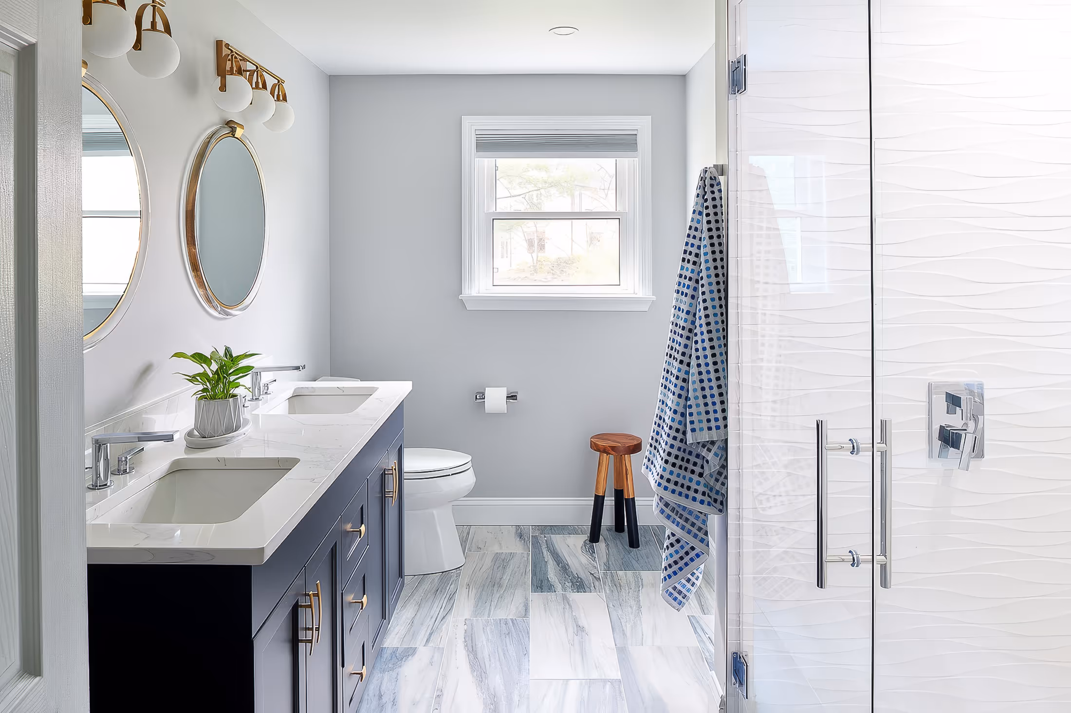 Modern bathroom with double sink vanity, round mirrors, white toilet, small wooden stool, and glass shower door with textured tiles.